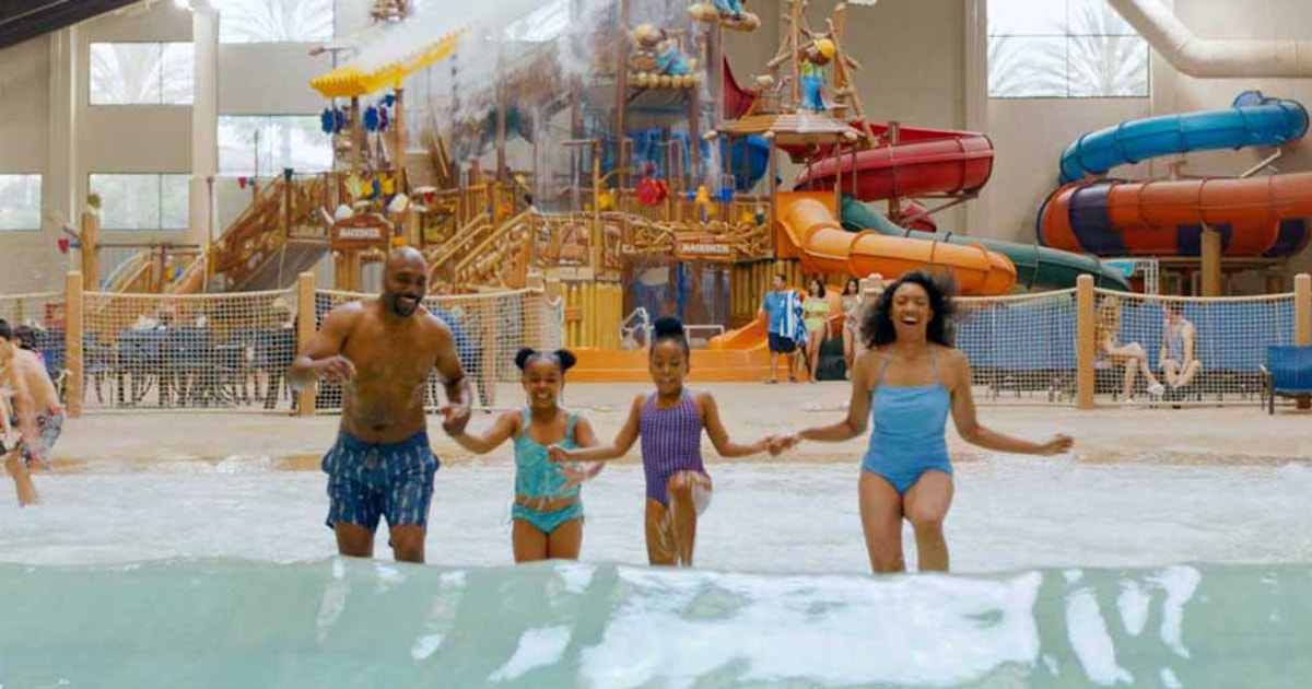 A family of four holds hands and smiles while walking through a large indoor wave pool at a water park.