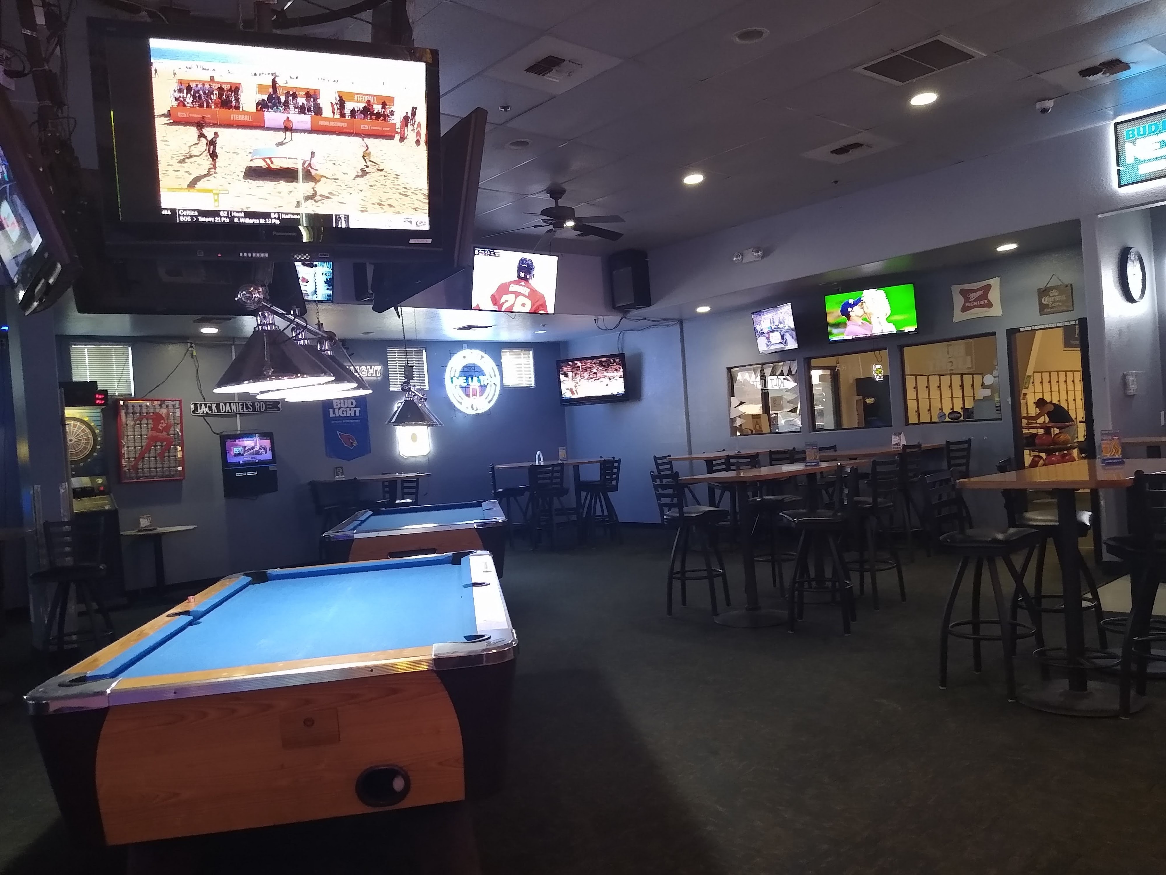 A dimly lit lounge with two billiard tables, high-top tables, and multiple televisions showing sports, with a glimpse of bowling lockers in the background.