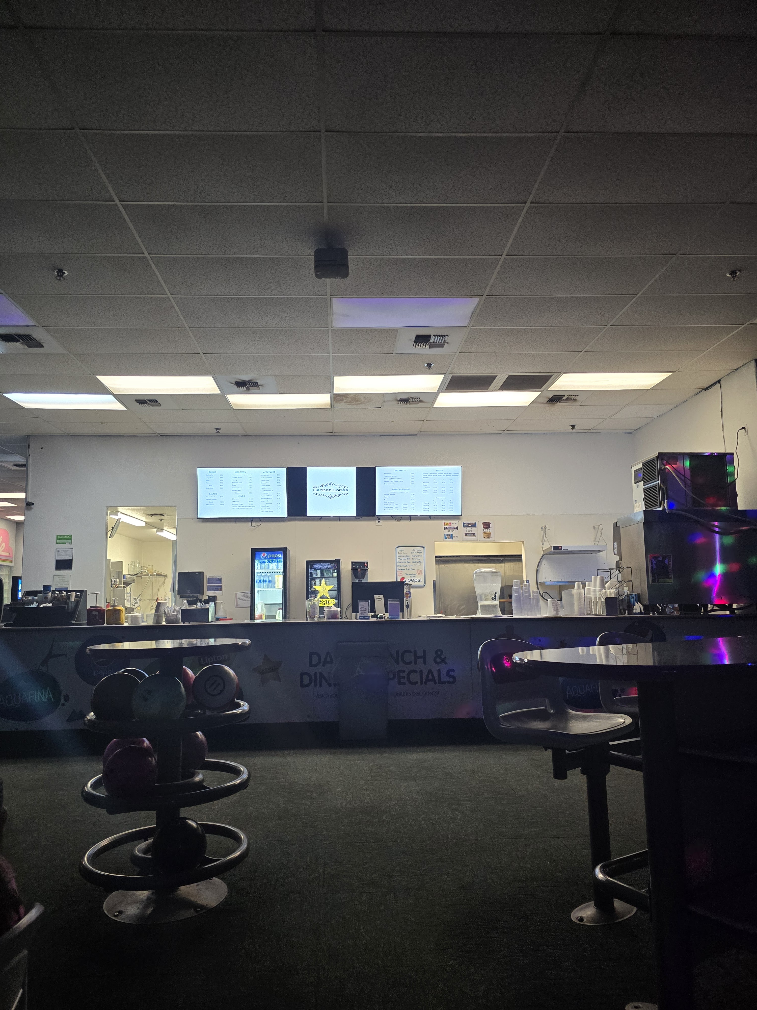 A dimly lit food service counter with digital menus and bowling balls on a rack in the foreground.