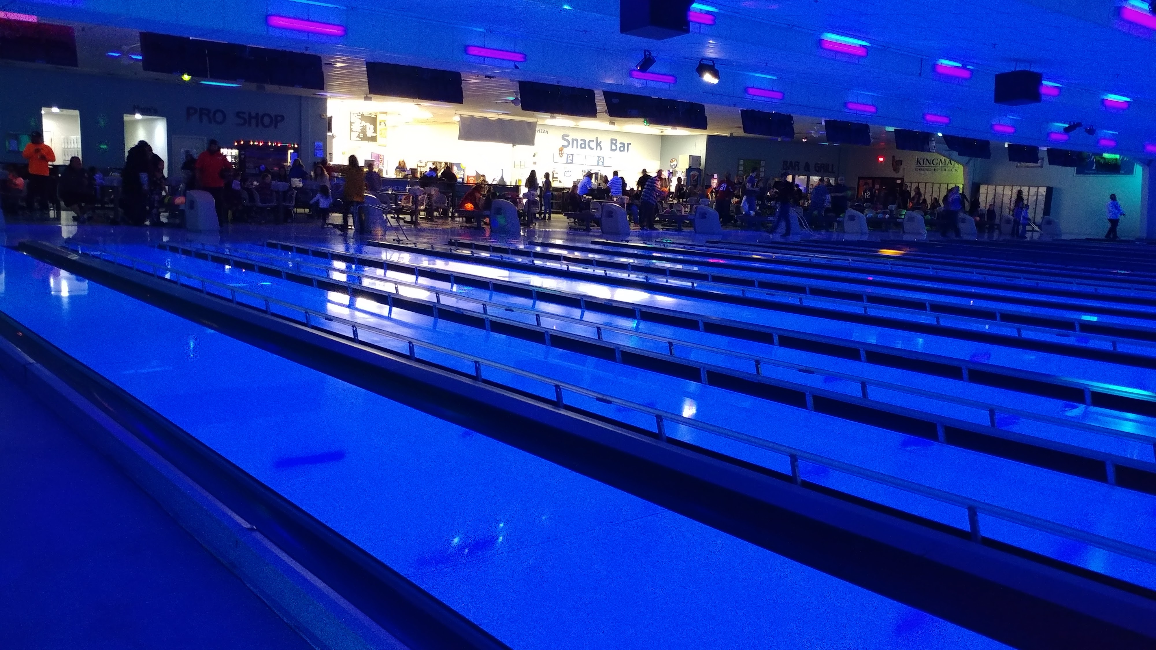 A wide view of a bowling alley illuminated by blue and purple cosmic lighting, with multiple lanes leading to a busy seating area, snack bar, and pro shop.