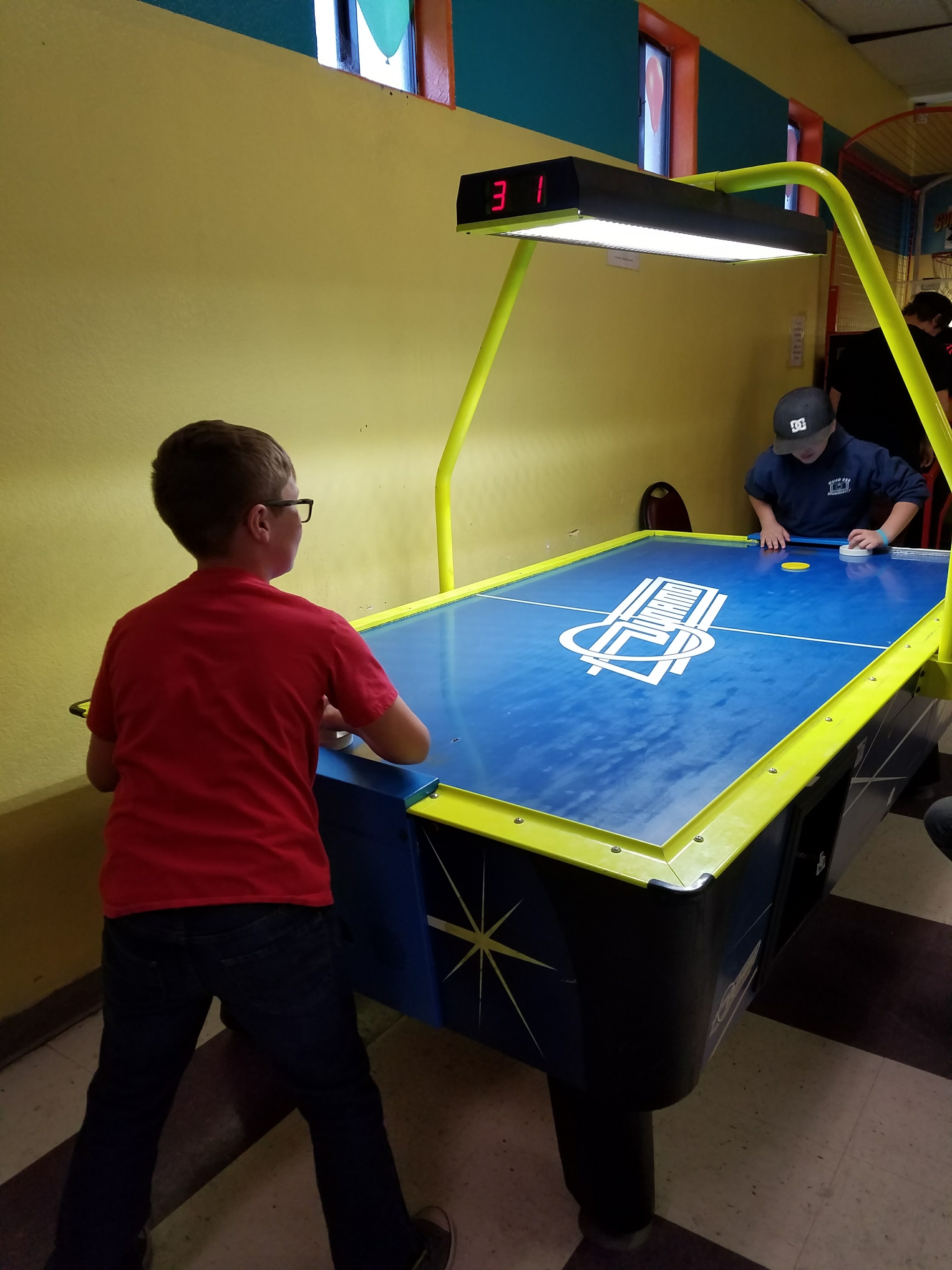 Two children play air hockey at a blue and yellow table under a brightly lit digital scoreboard.
