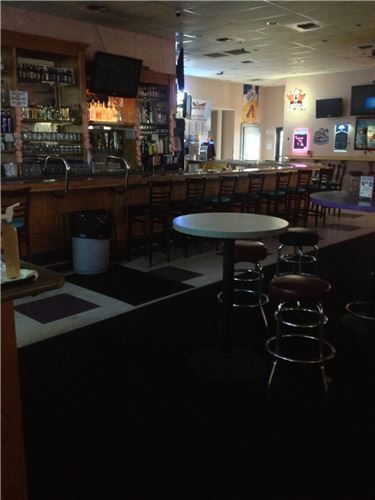 An empty bar and lounge area with a long wooden counter, bar stools, high-top tables, and shelves of bottles.