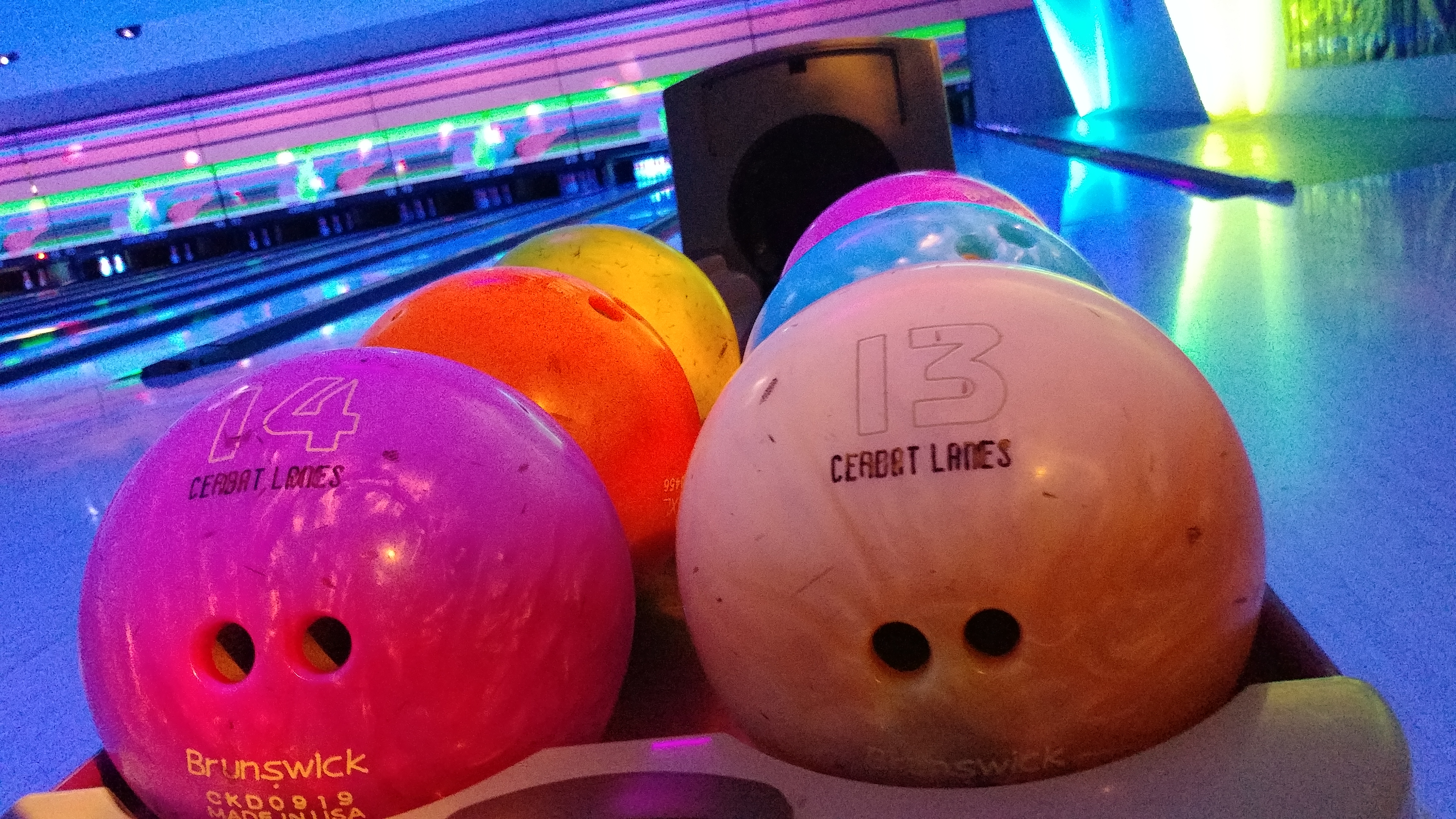 Vibrant pink, orange, and white house bowling balls on a rack with neon-lit lanes in the background.
