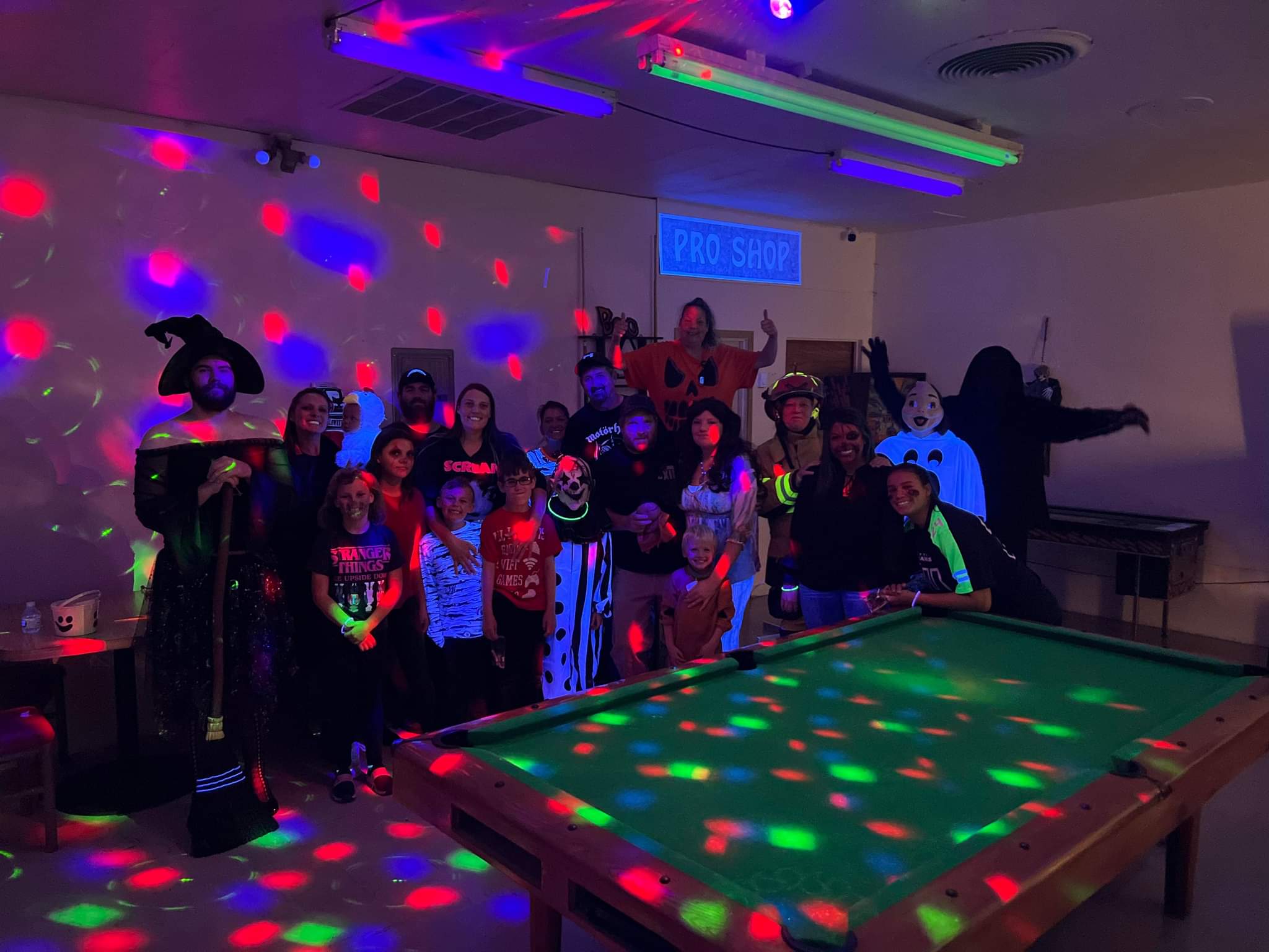 A large group of people in Halloween costumes poses behind a pool table under colorful disco lights.