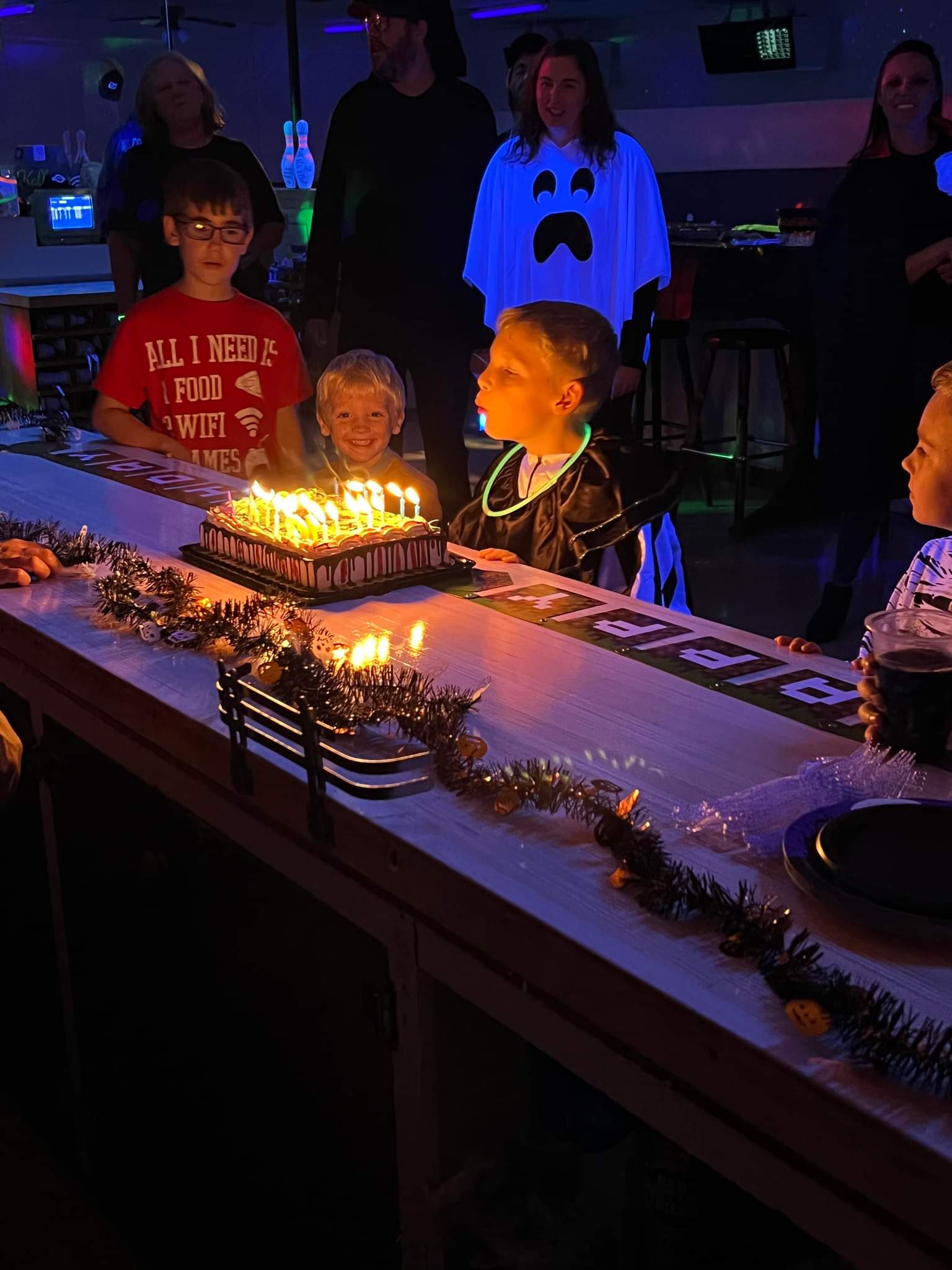 A group of children and adults at a bowling alley gather around a birthday cake with lit candles.