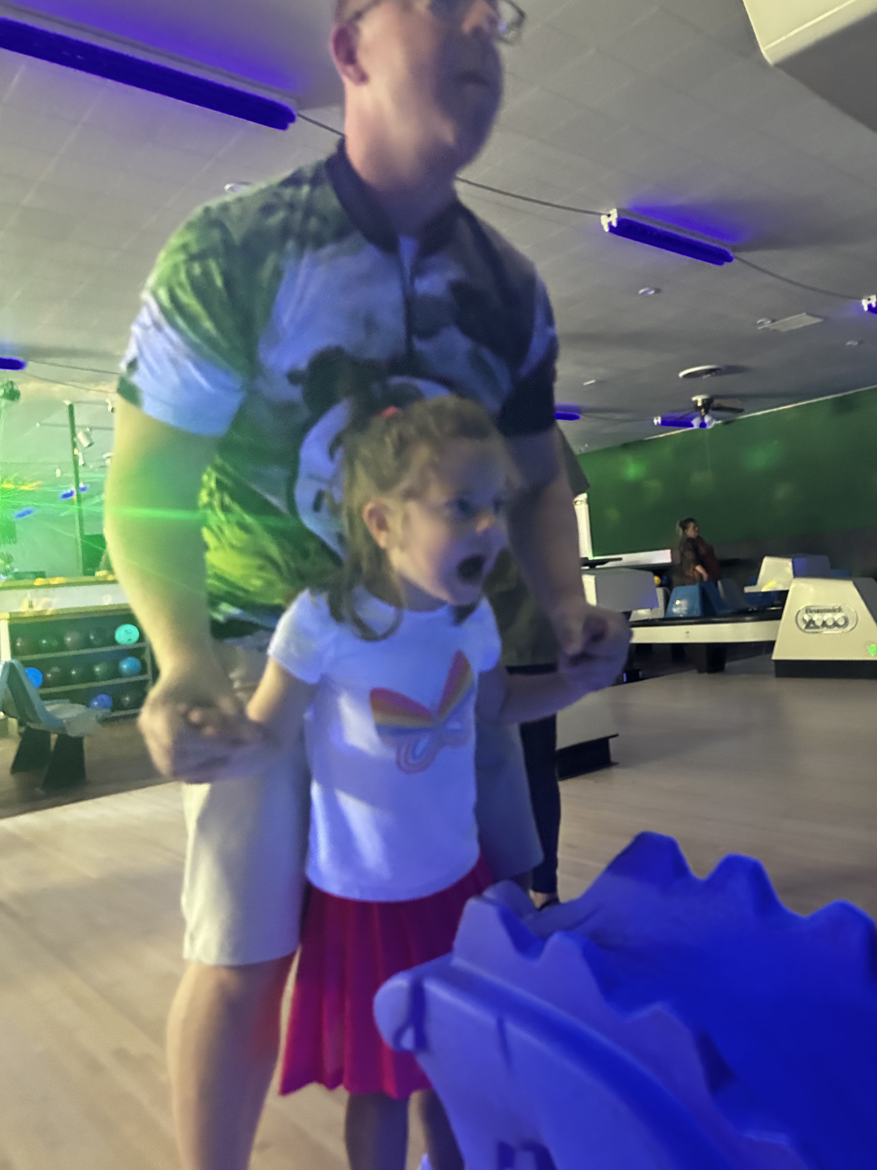 A young girl reacts with excitement while using a blue bowling ramp during a cosmic bowling session with a man.
