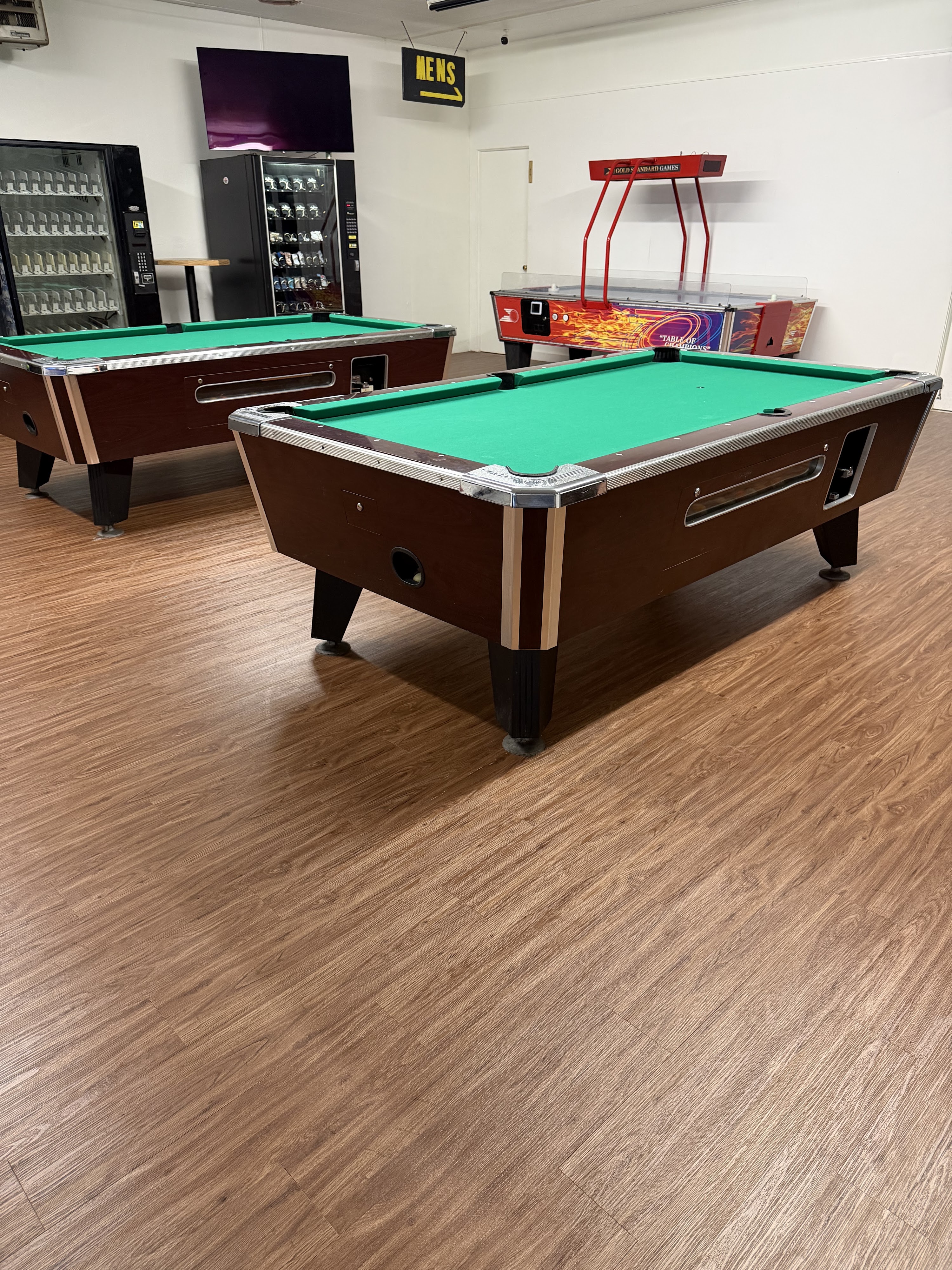 Two green felt pool tables and a red air hockey table in a brightly lit game room with wood-style flooring.
