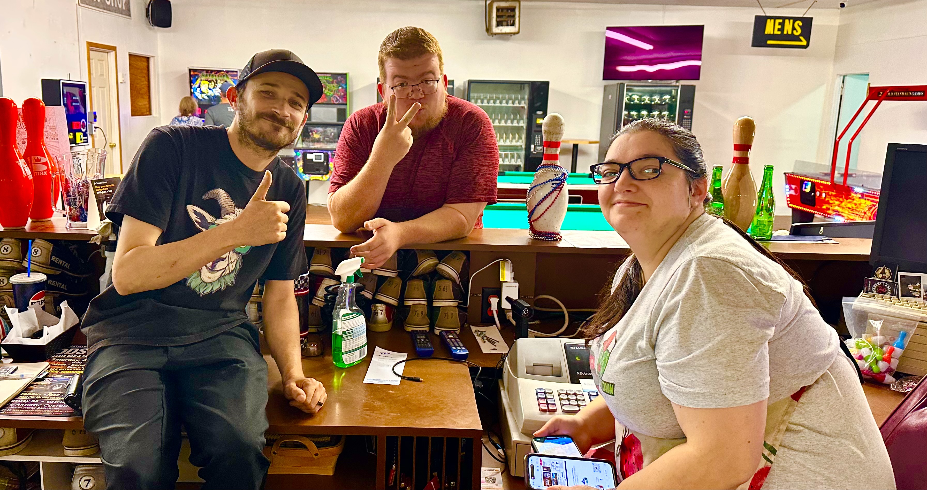 Three people posing behind a bowling alley service counter with rental shoes and arcade games visible in the background.