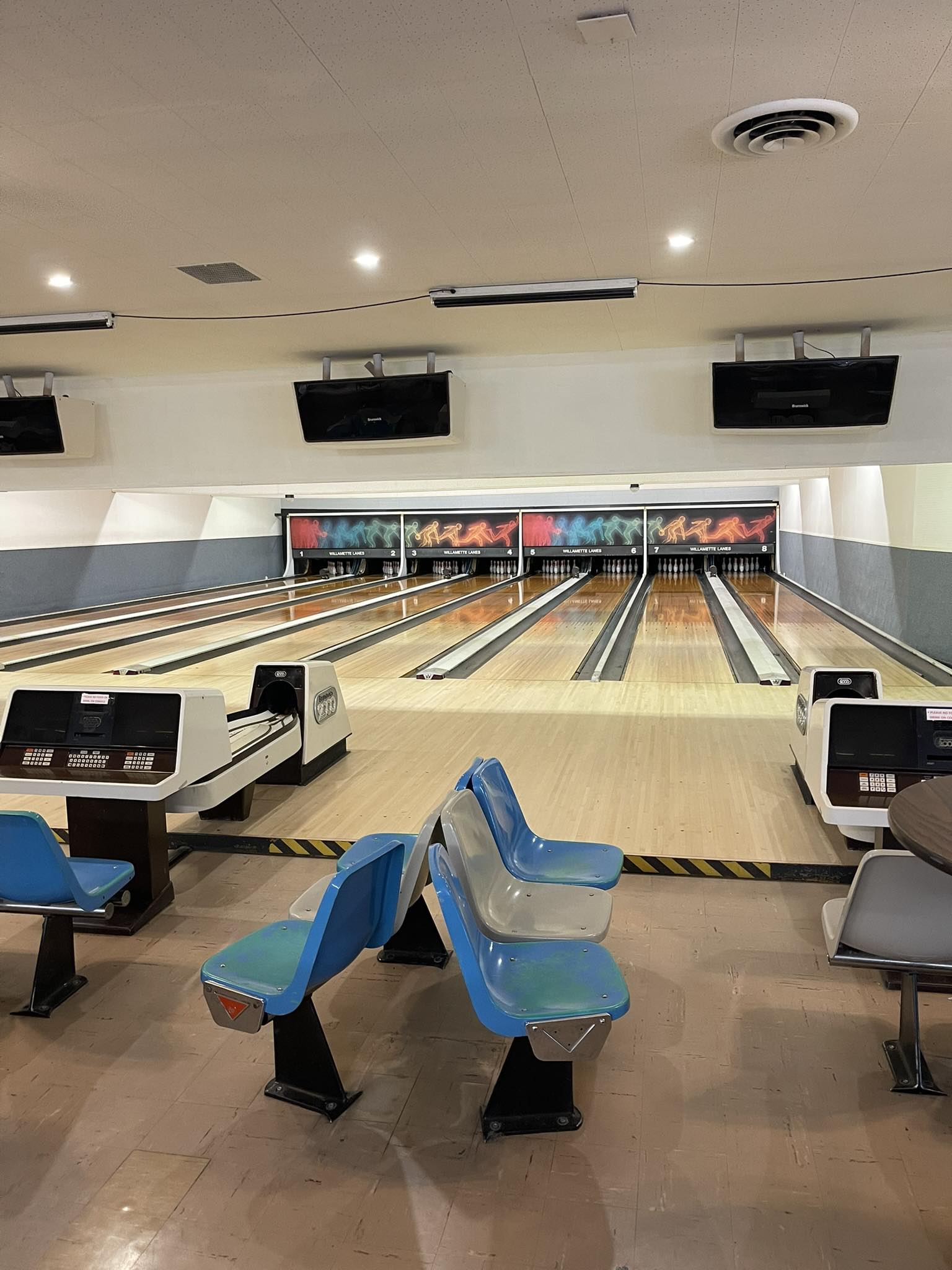 An interior view of eight wooden bowling lanes with blue and gray plastic seats in the foreground.