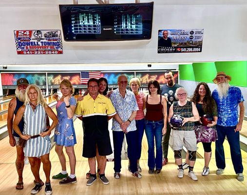 A diverse group of eleven people posing together on bowling lanes under a scoring monitor.