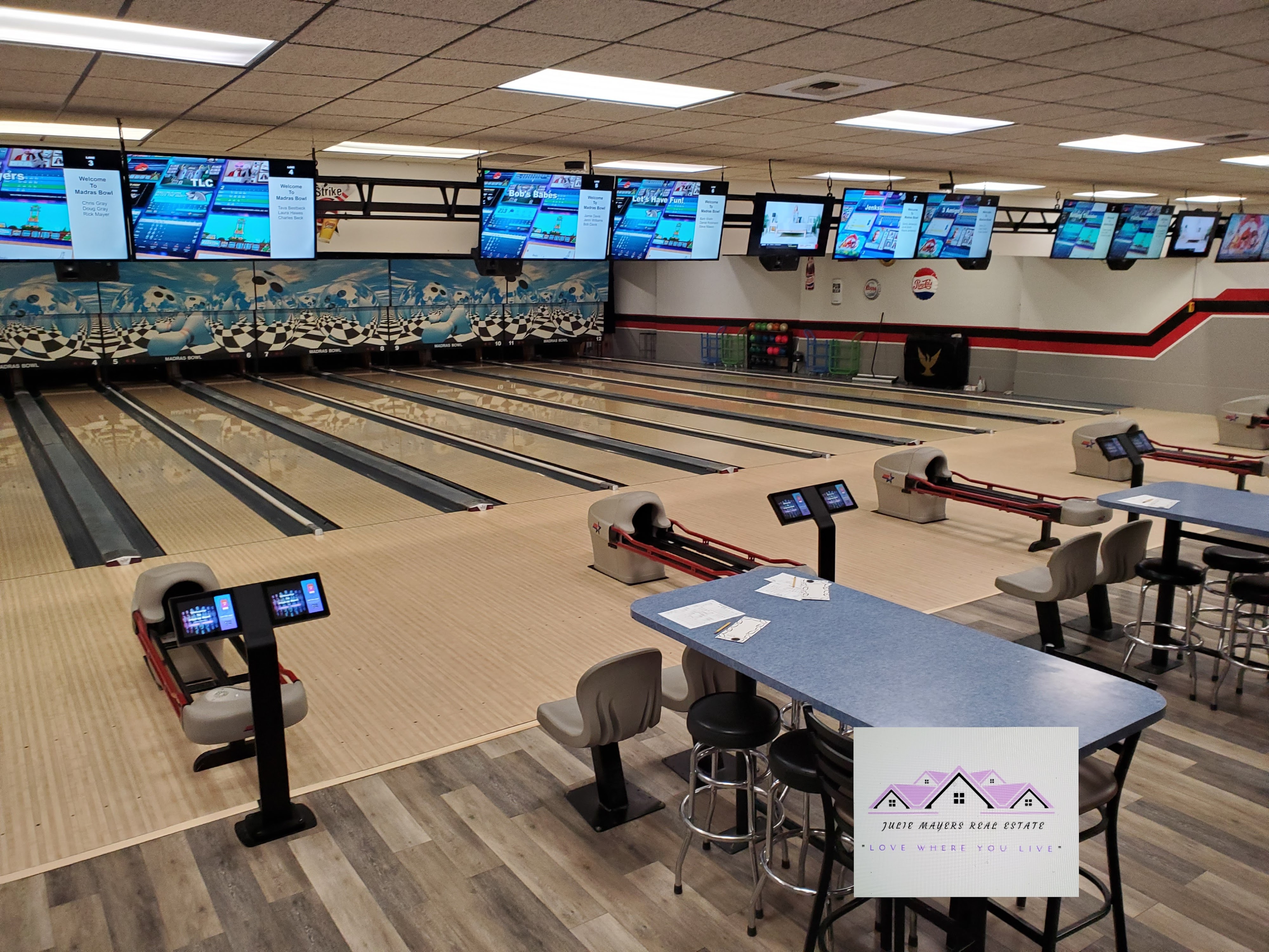 Wide interior view of a bowling alley with multiple lanes, scoring monitors, and blue high-top tables with stools.