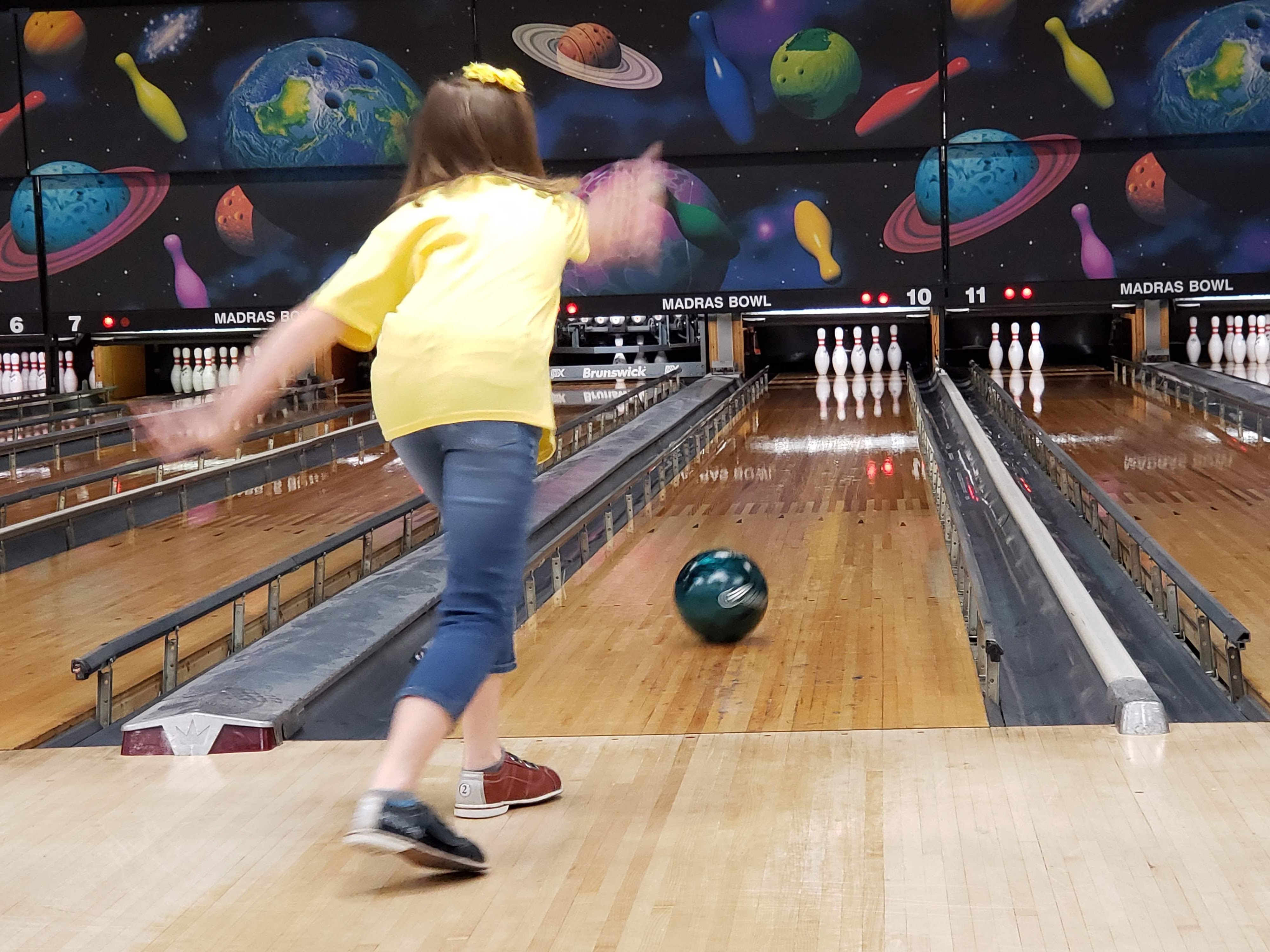 A young girl in a yellow shirt watches her bowling ball roll down a lane with bumpers.