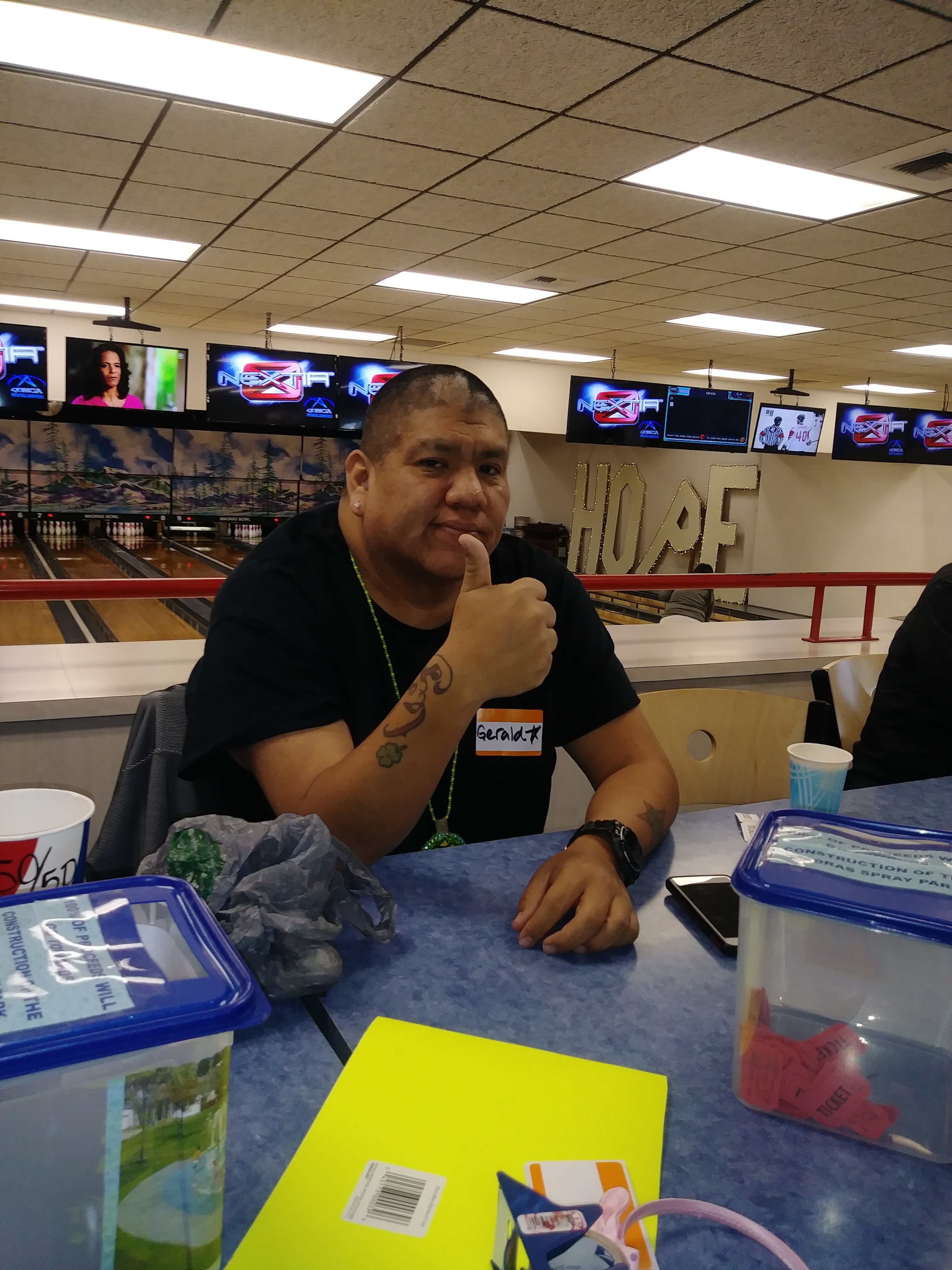 A man with a name tag sits at a table and gives a thumbs up inside a bowling alley.