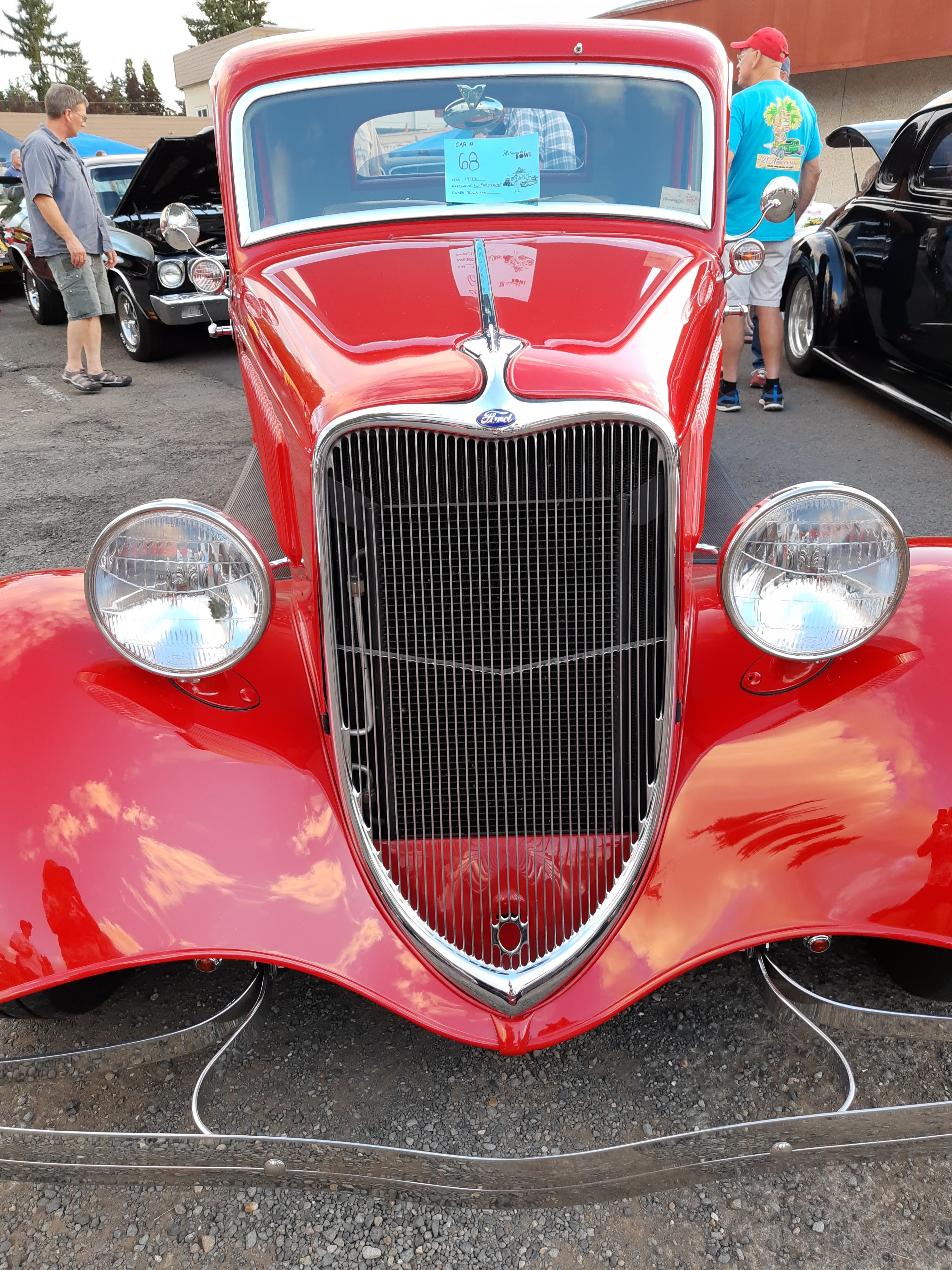 Close-up front view of a vibrant red vintage Ford coupe at a car show in a parking lot.