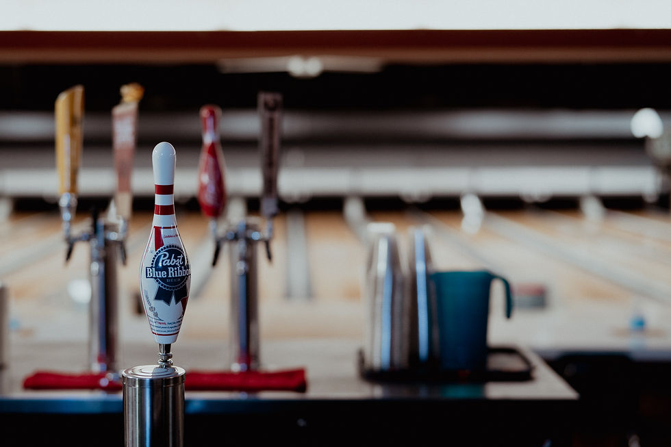 A bowling-pin-shaped beer tap handle at a bar with blurred bowling lanes in the background.