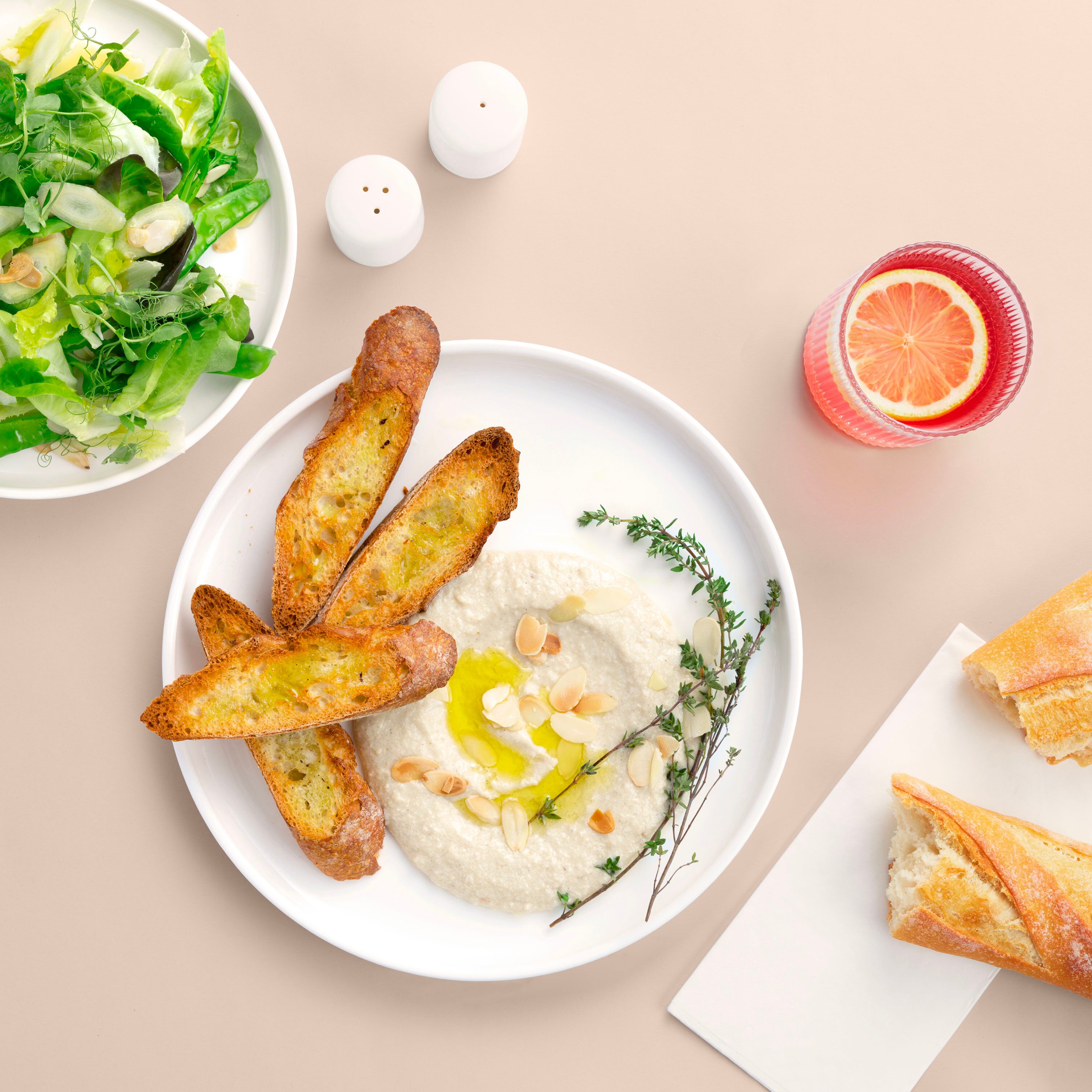 A top-down view of a food spread featuring a dip with toasted bread, a green salad, and a pink citrus drink.