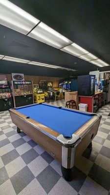 A blue felt billiard table on a checkered floor in an arcade room with several gaming machines in the background.