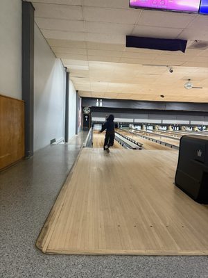 A person in a dark hoodie stands on a wood bowling lane approach facing the pins.