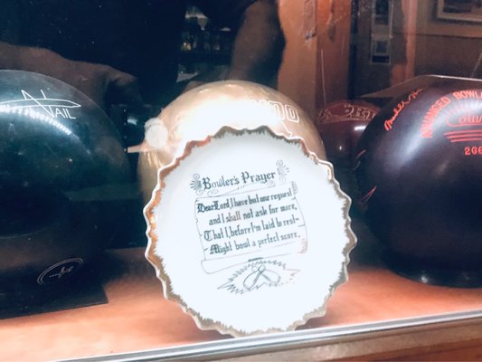 A decorative scalloped plate featuring the Bowler's Prayer displayed in front of various bowling balls.