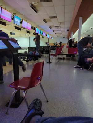 A bowling alley interior with red chairs, scoring consoles, and people bowling under overhead monitors.