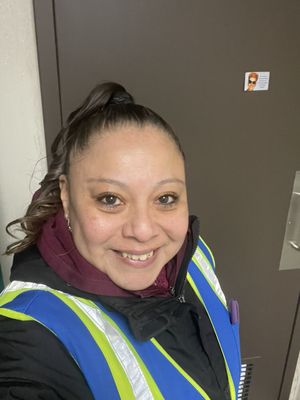 A smiling woman wearing a blue and yellow high-visibility safety vest over a black jacket.