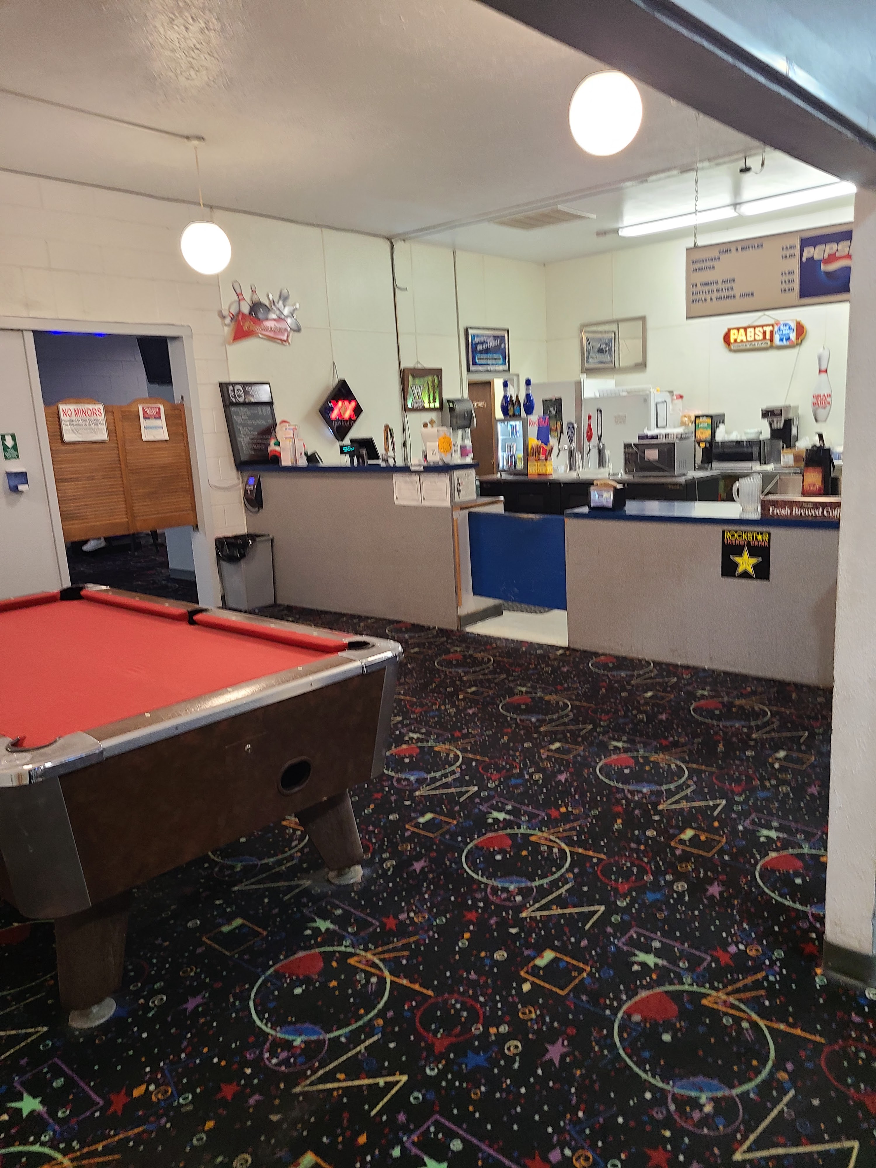 A billiards table with red felt sits on a patterned carpet in front of a snack bar counter.
