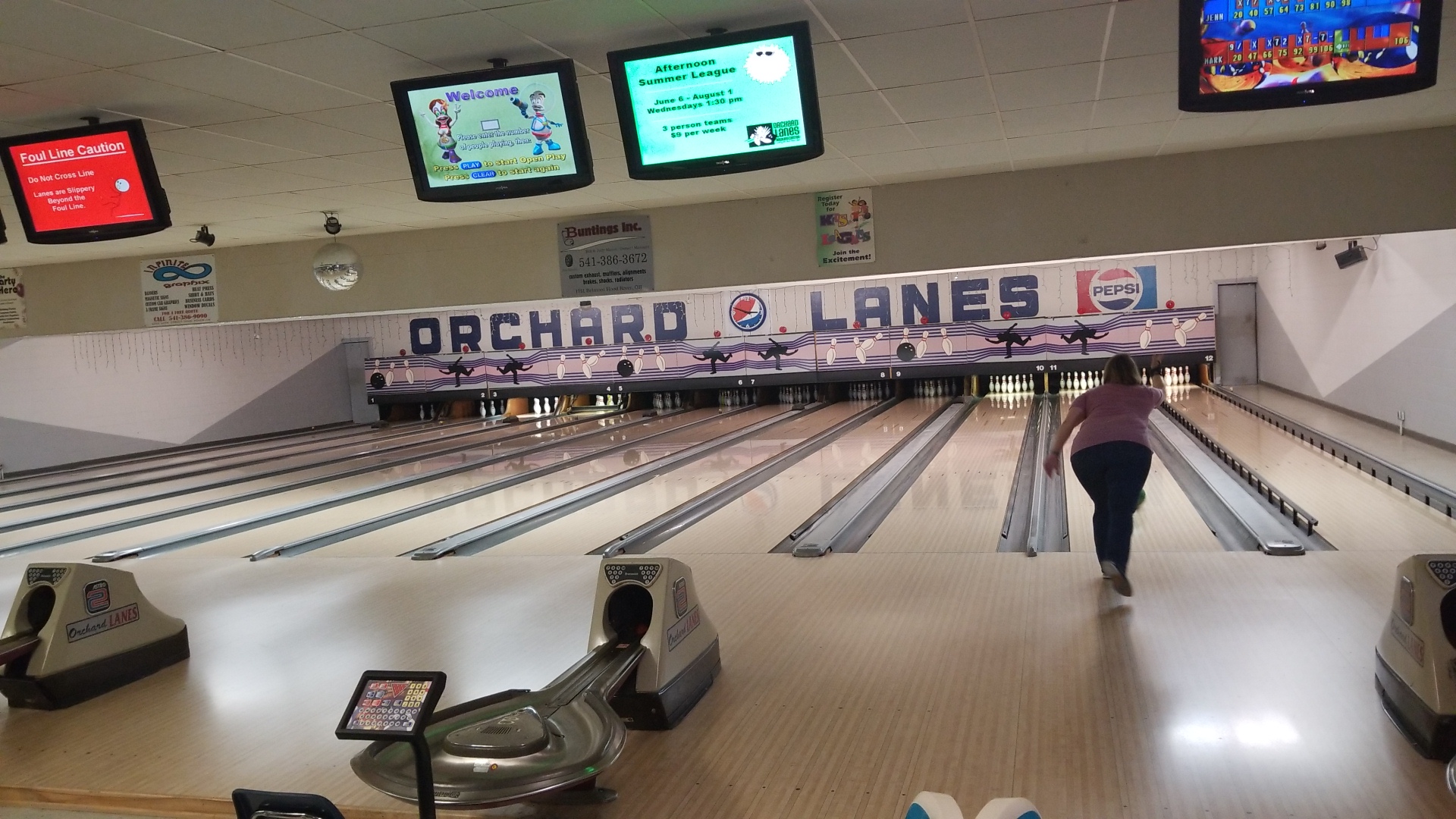 A woman bowls at Orchard Lanes under overhead scoring monitors and a large center logo.