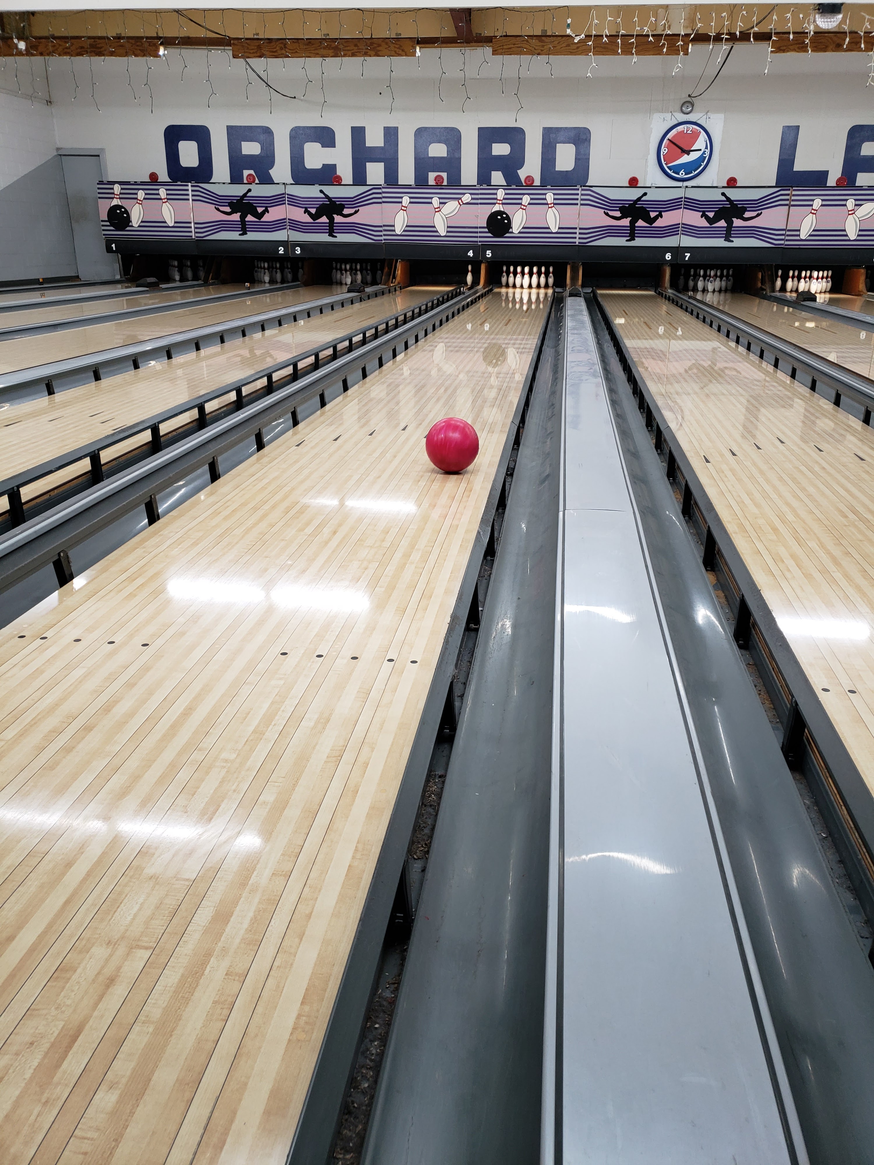 A red bowling ball rolls down a wooden lane towards pins at Orchard Lanes.
