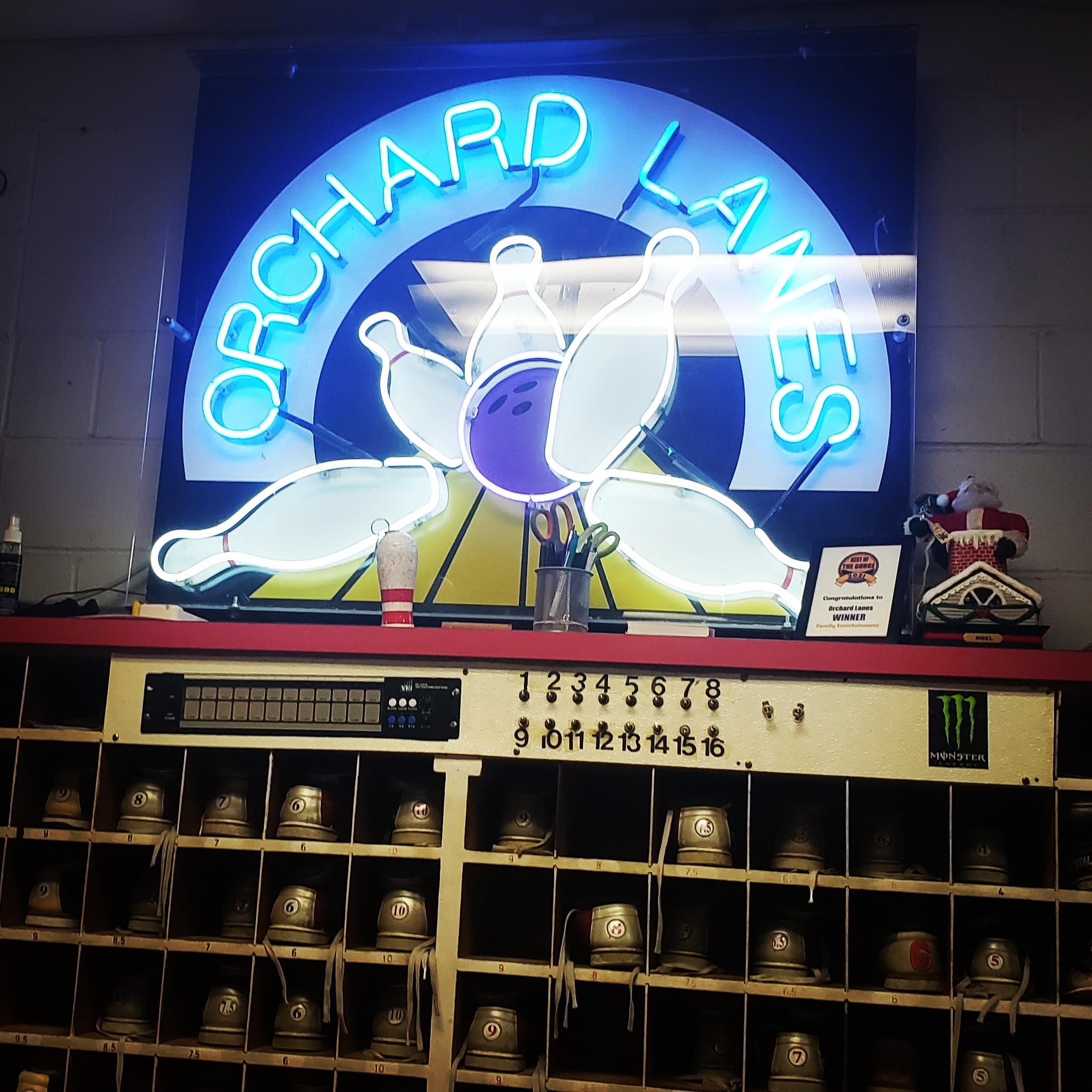 A blue neon Orchard Lanes sign sits on a shelf above a wooden rack full of rental bowling shoes.