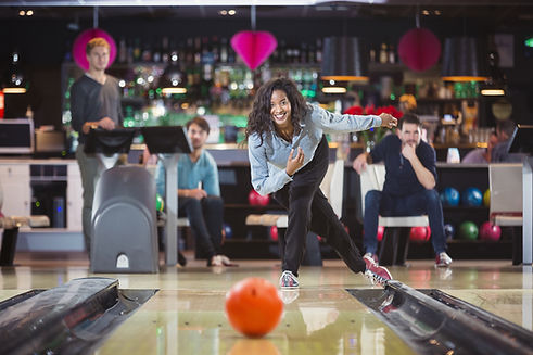 A woman bowls a ball down the lane while friends watch from the seating area at a bowling center.