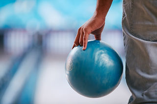 Close-up of a person's hand holding a blue bowling ball with a blurred bowling lane in the background.