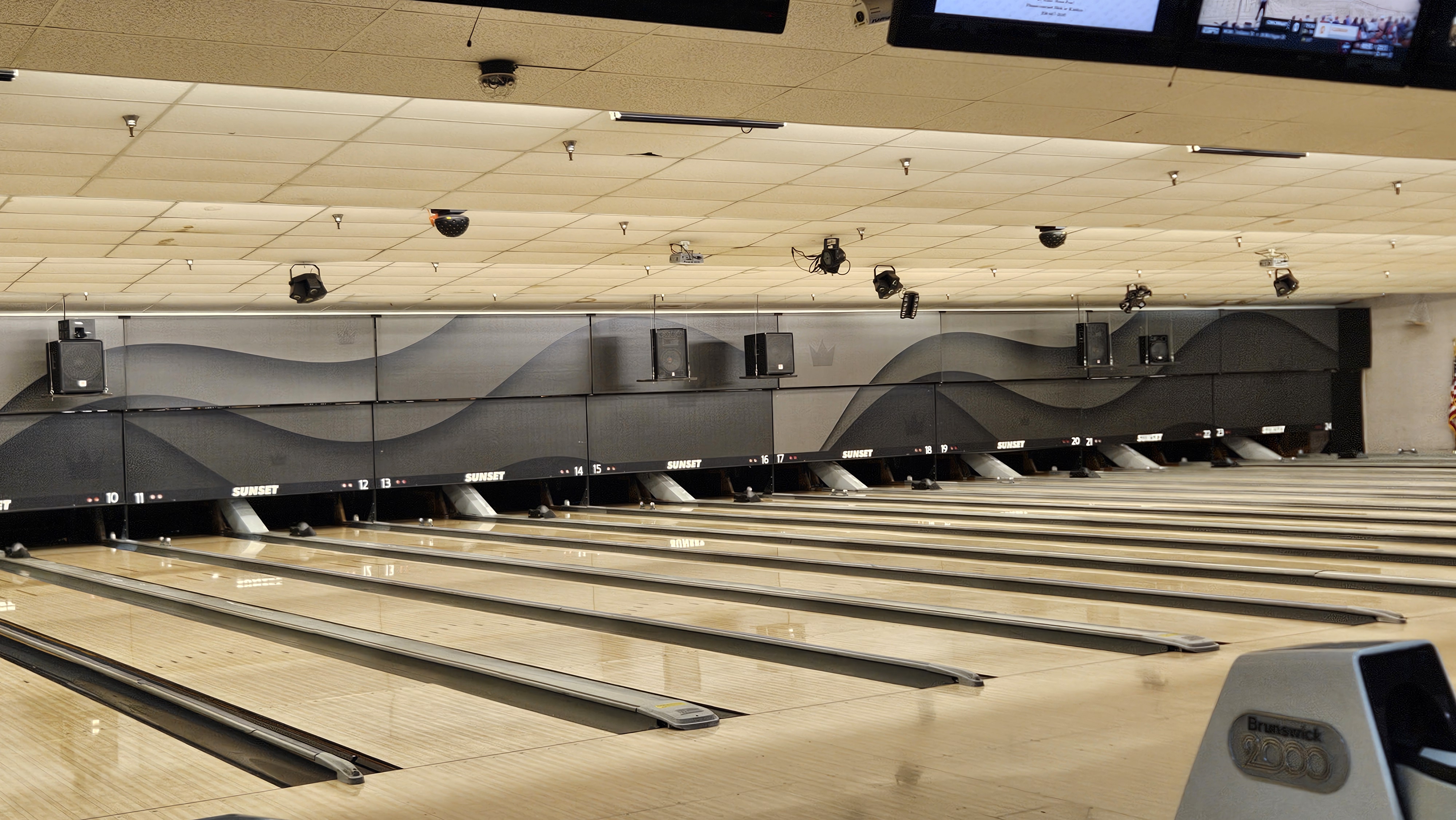 Wide view of polished wooden bowling lanes with grey wave-patterned backboards and overhead monitors.
