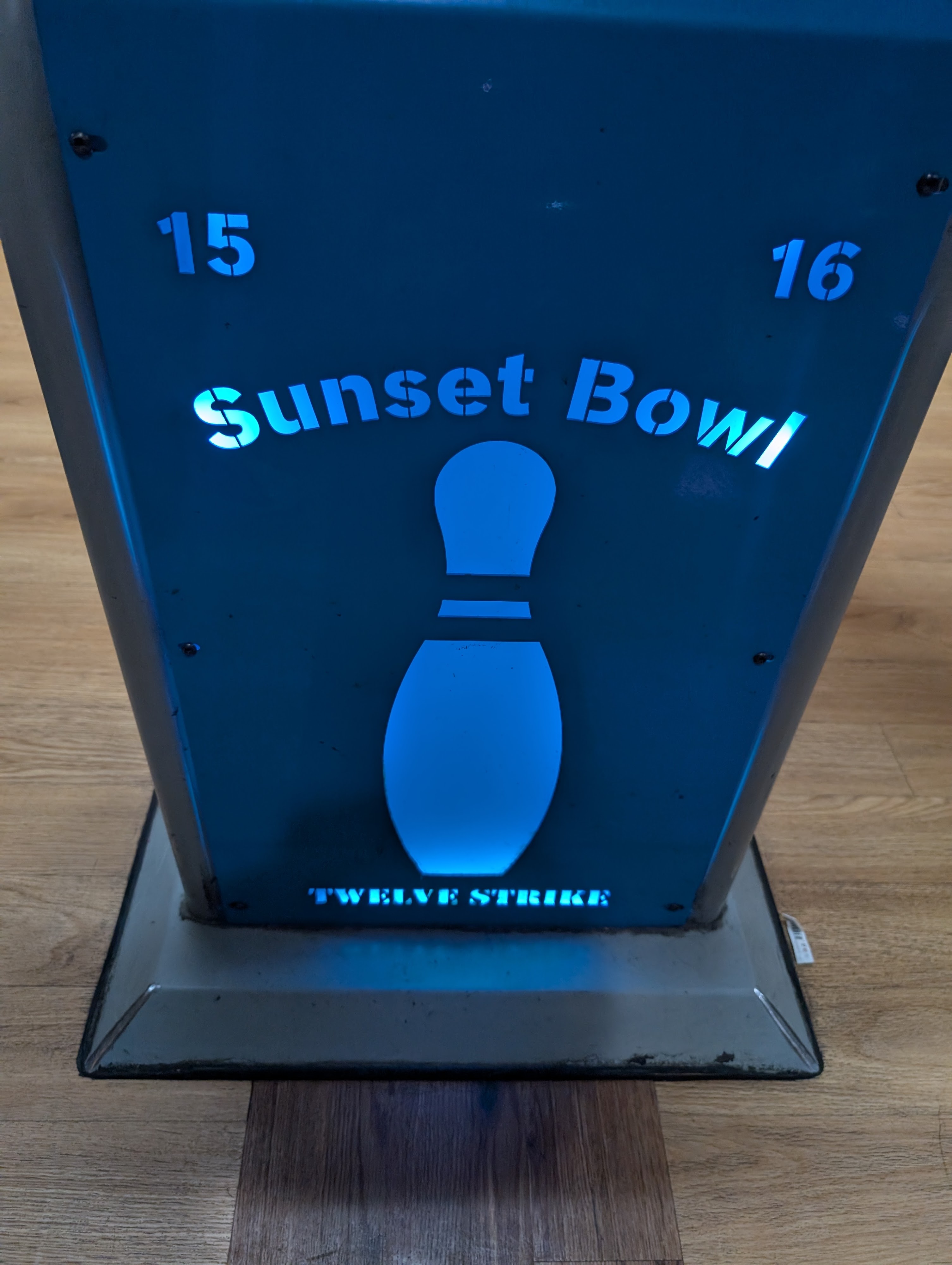 A blue backlit metal pedestal featuring a bowling pin cutout and the words Sunset Bowl between lanes 15 and 16.