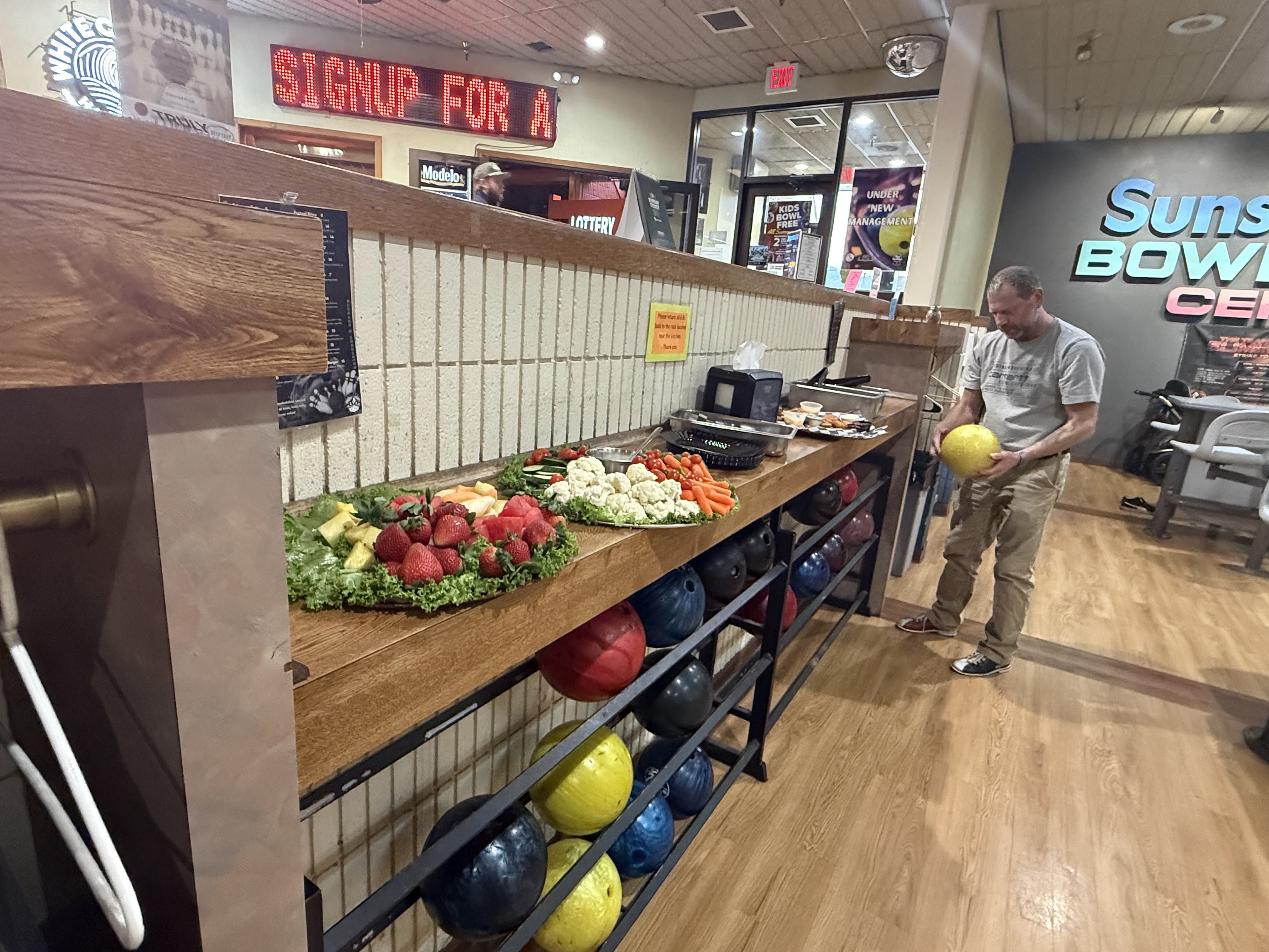 Trays of fresh fruit and vegetables on a wooden counter above racks of colorful bowling balls.