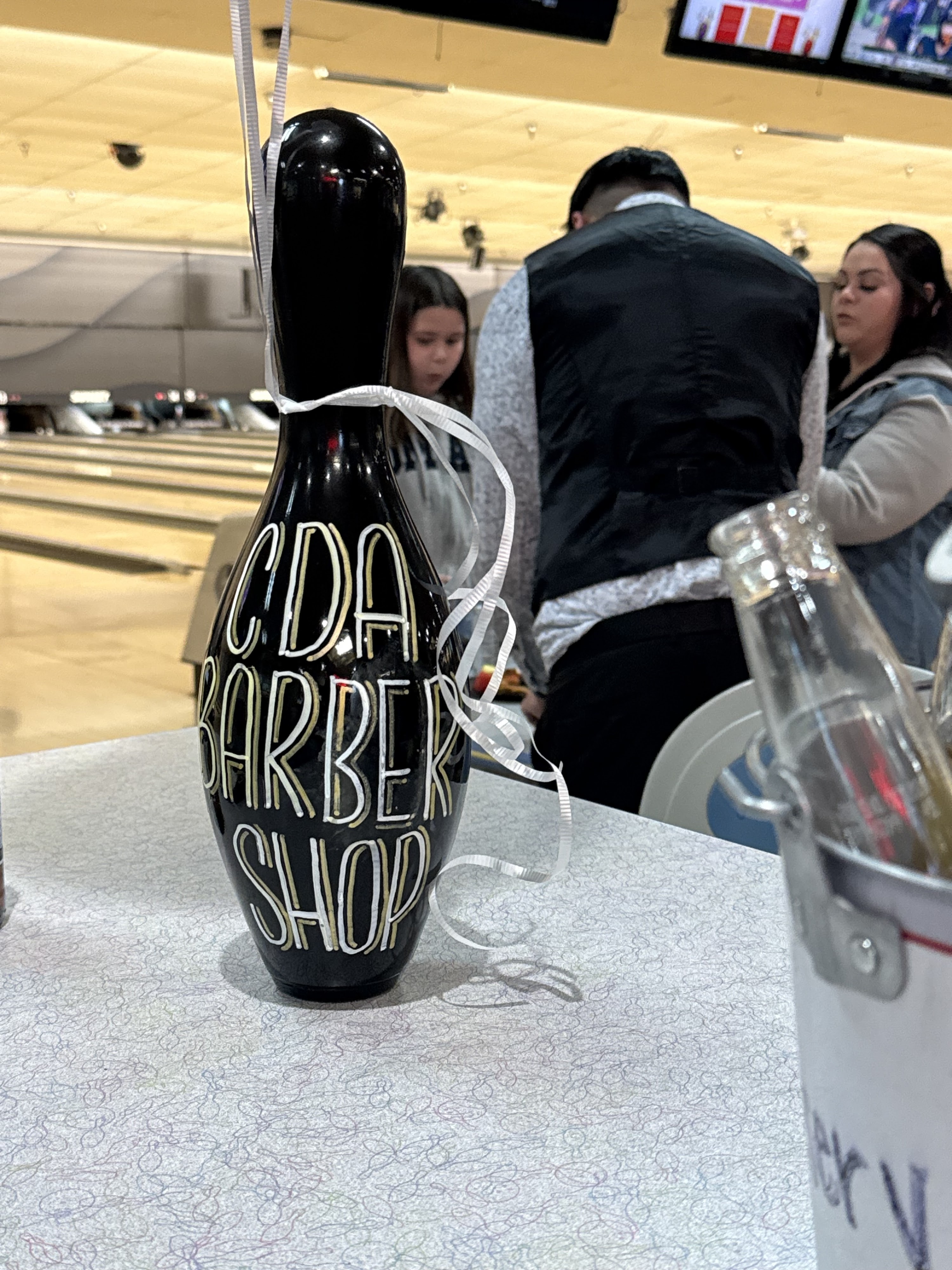 A black bowling pin with CDA Barber Shop hand-painted on it sits on a table inside a bowling center.