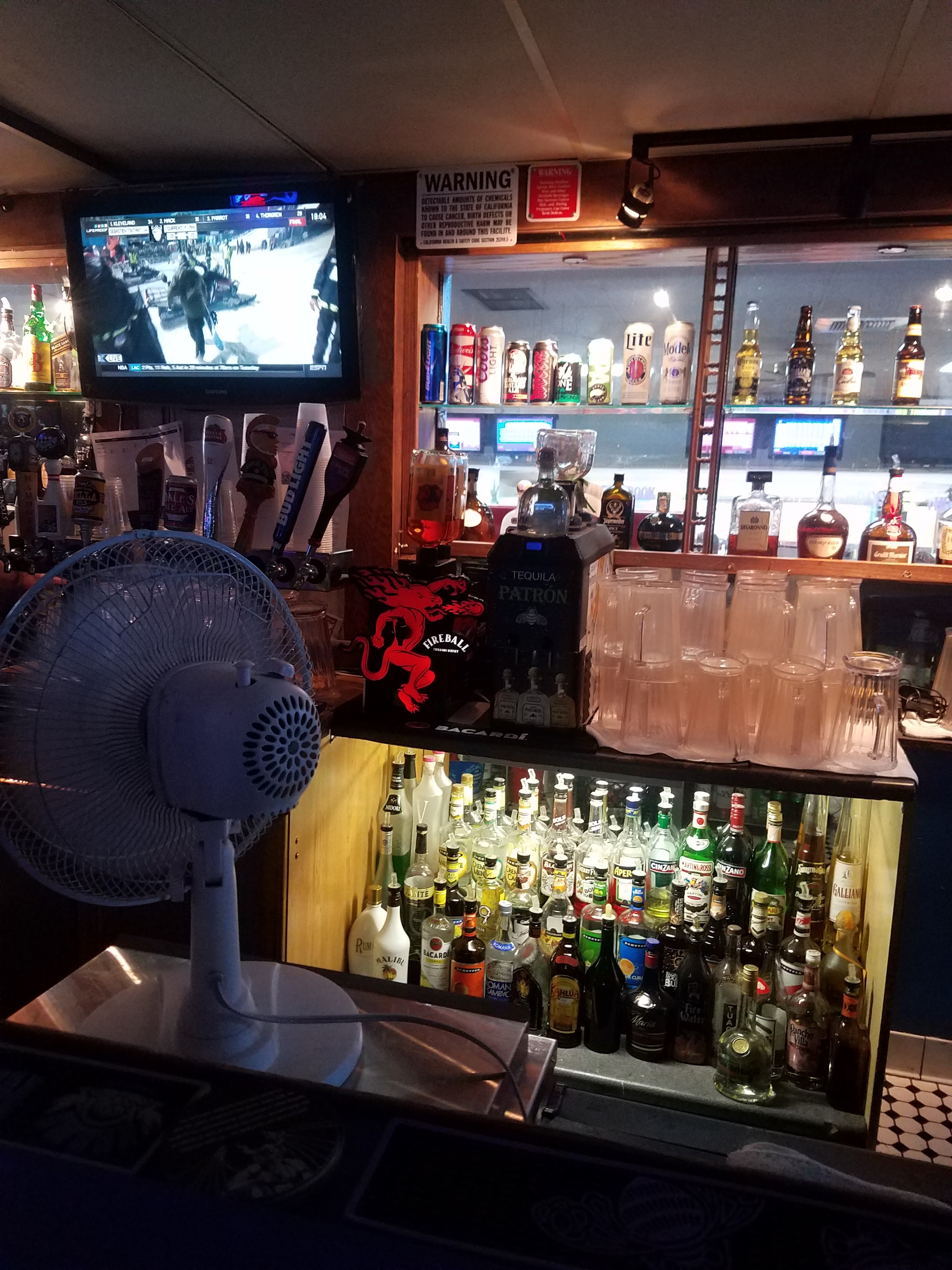 A well-stocked bowling alley bar with various liquor bottles, beer taps, and a television showing sports.