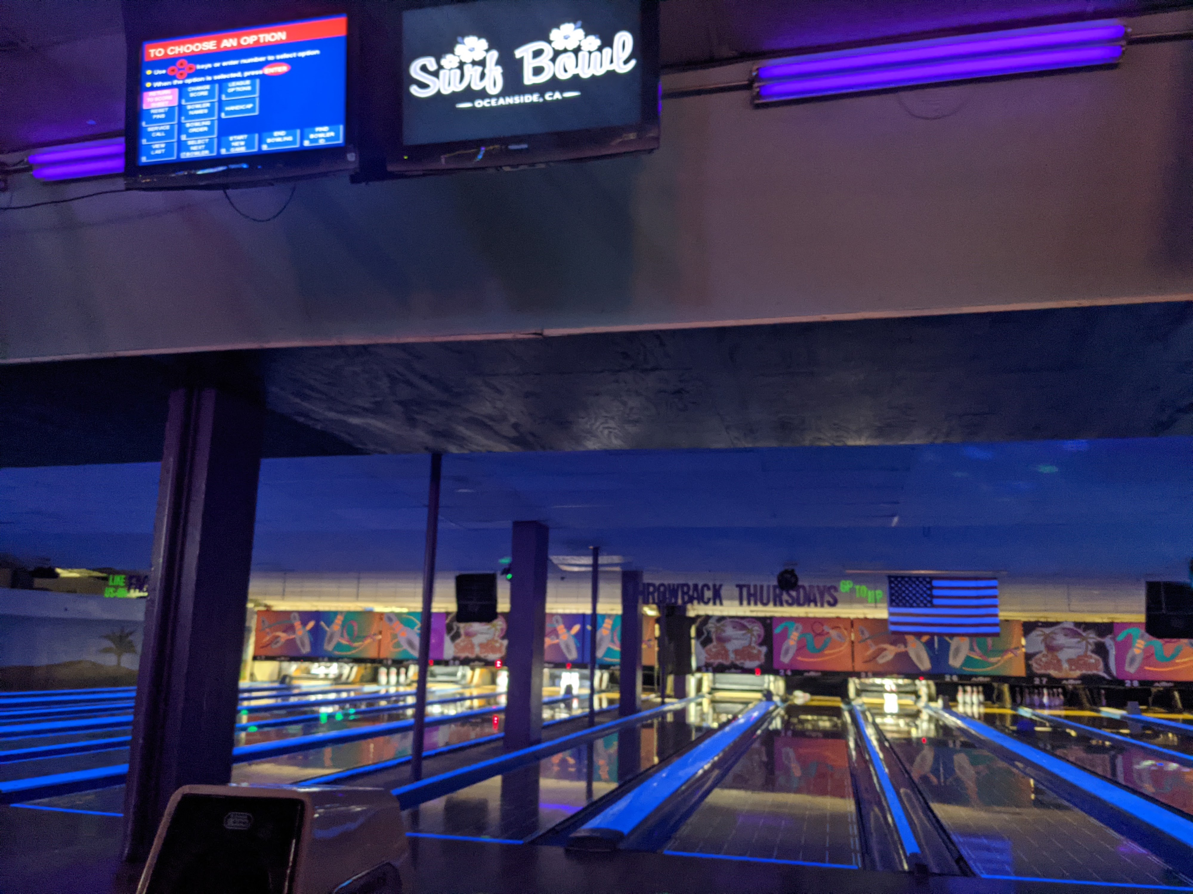 Several bowling lanes illuminated with blue cosmic lighting under a monitor displaying the Surf Bowl logo.