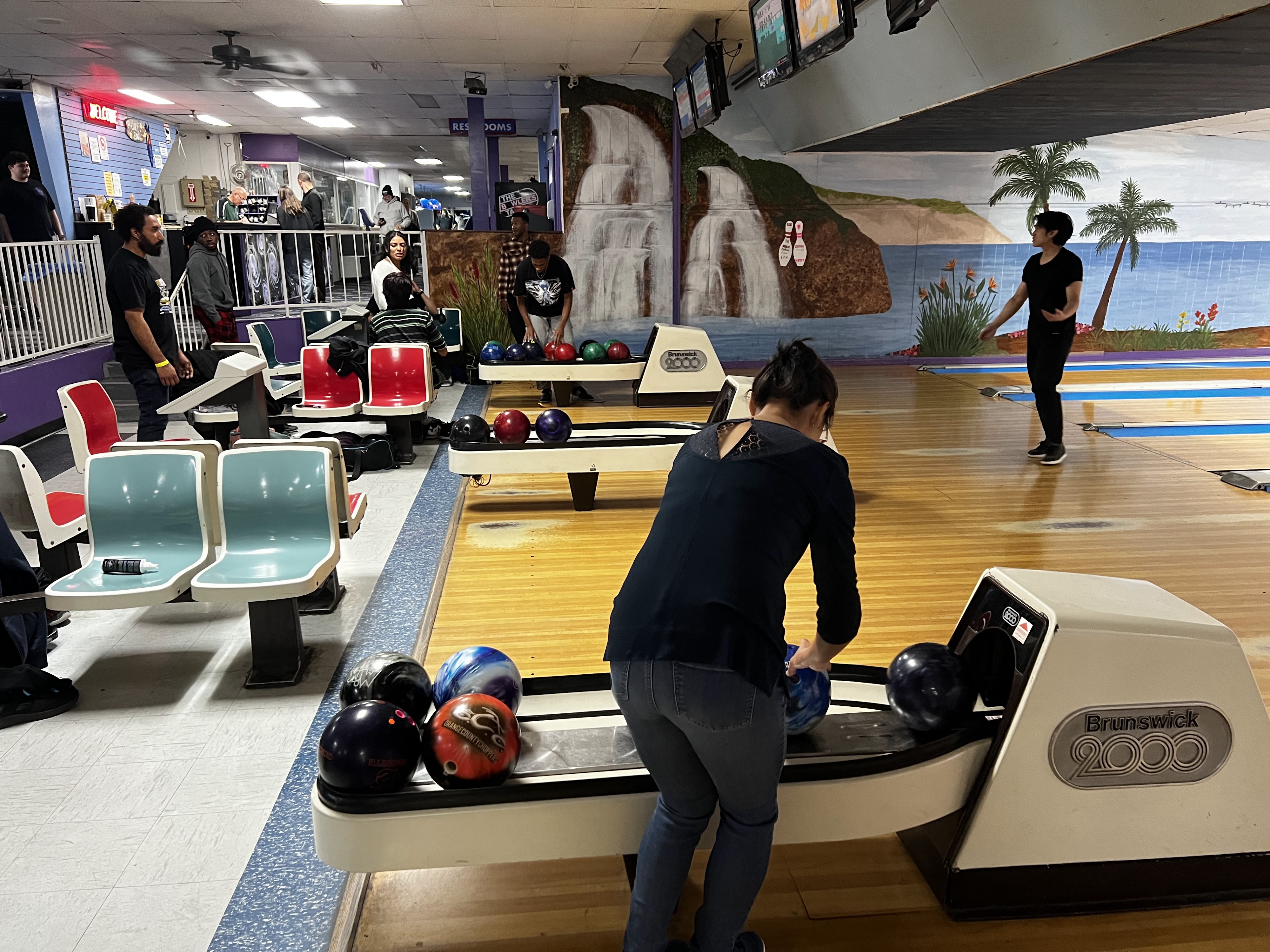 People bowling at a center with wooden lanes, colorful retro seating, and tropical beach murals on the walls.