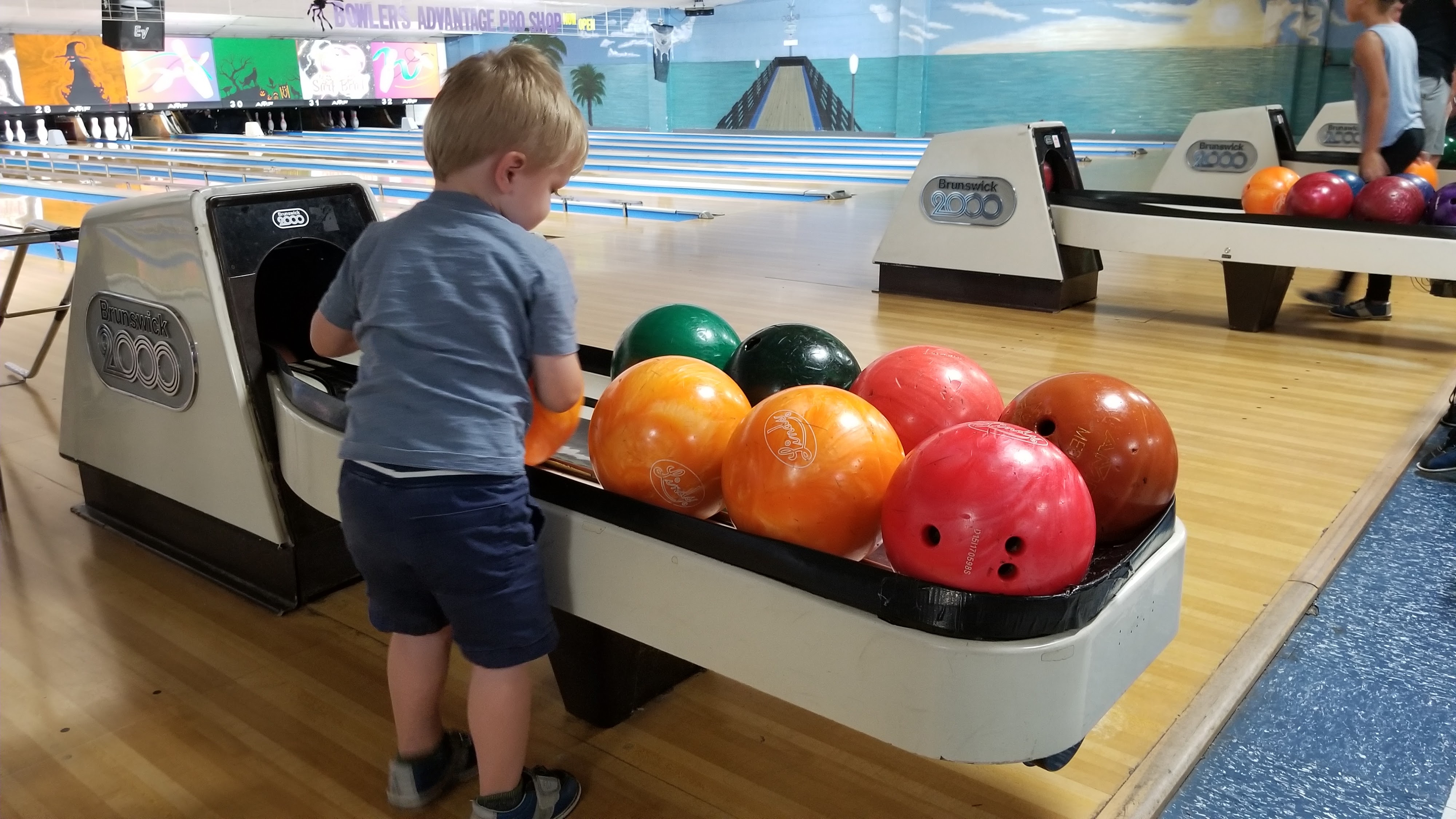 A young boy reaches for a colorful bowling ball at the ball return in a brightly lit bowling center.