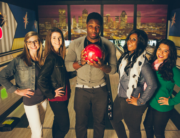 A group of five smiling young adults pose together on a bowling lane, with the center man holding a red bowling ball.