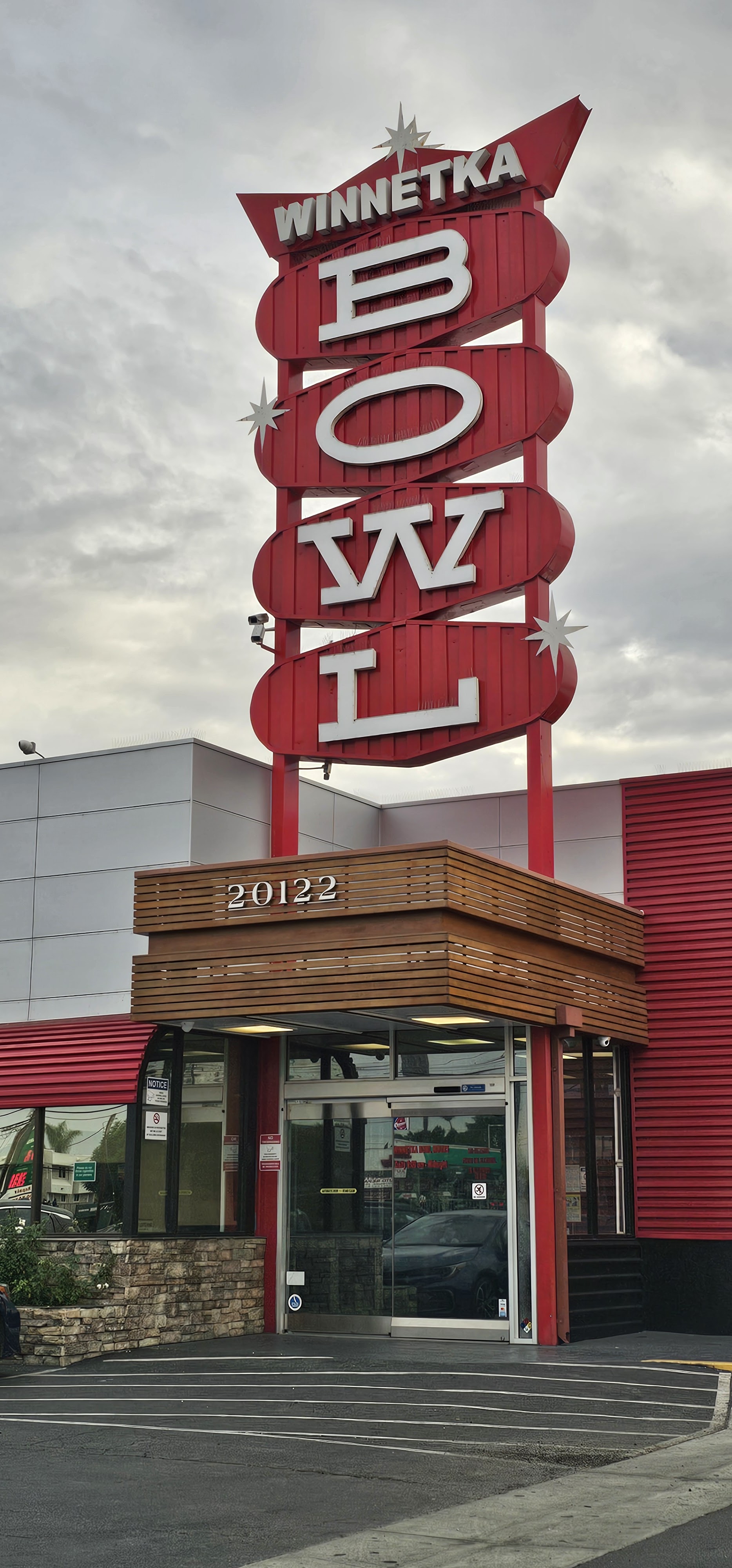 A tall red vintage Winnetka Bowl sign stands above a modern glass entrance under a cloudy sky.