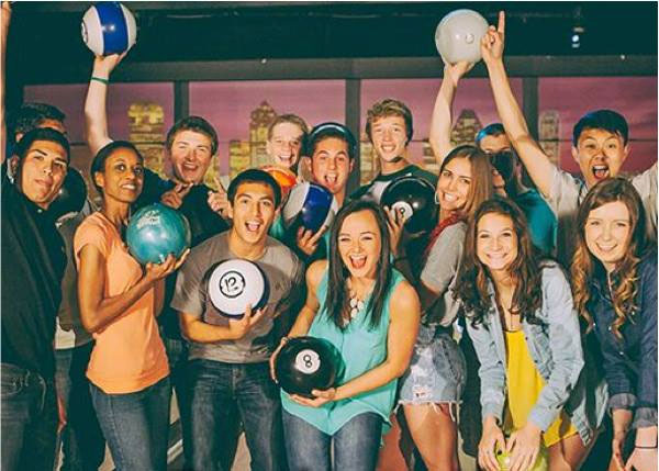 A large group of smiling young adults holding bowling balls and cheering together at a bowling center.