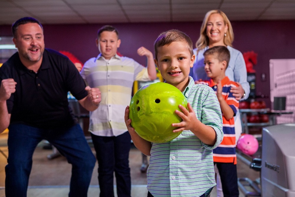 A young boy holds a green bowling ball while his family cheers behind him at a bowling alley.