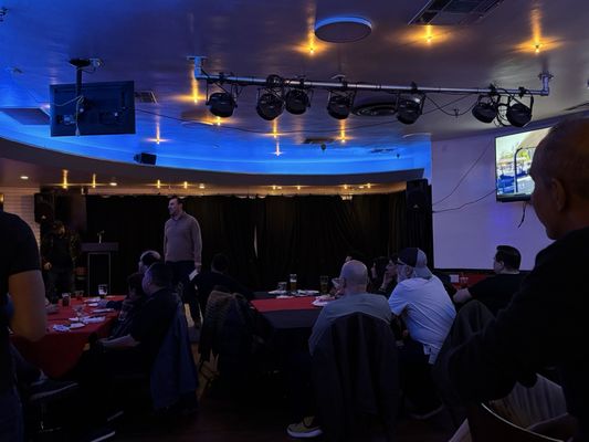 A group of people seated at banquet tables in a dimly lit room with blue ceiling lights watching a presentation.