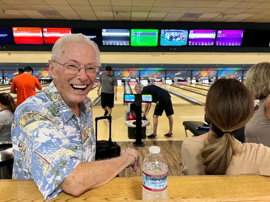 A smiling man in a Hawaiian shirt poses at a bowling alley with lanes and score monitors in the background.