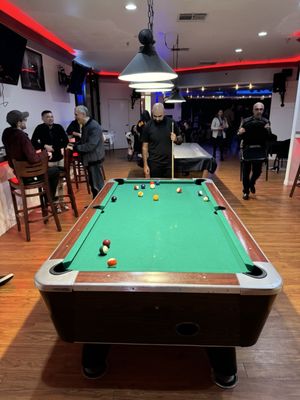 A man holding a cue stick stands at a green pool table in a lounge area with people in the background.