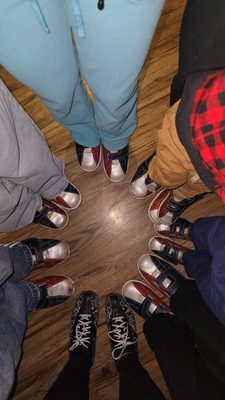 A circle of people seen from above, all wearing rental bowling shoes on a wooden floor.
