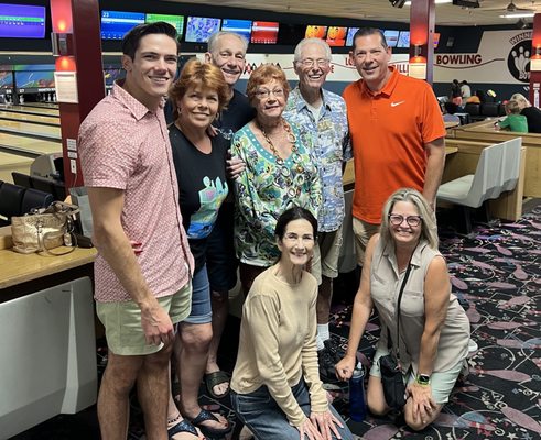 A group of seven smiling people posing for a photo together at a bowling center with lanes in the background.