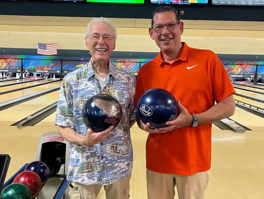 Two men smile while holding bowling balls at a bowling alley with lanes in the background.