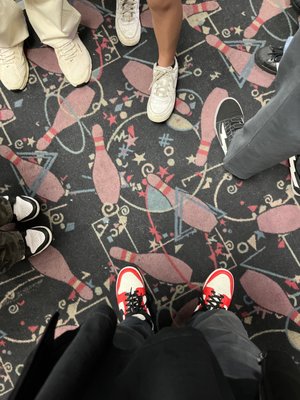 Top-down view of several people's sneakers standing on a carpet patterned with bowling pins and stars.